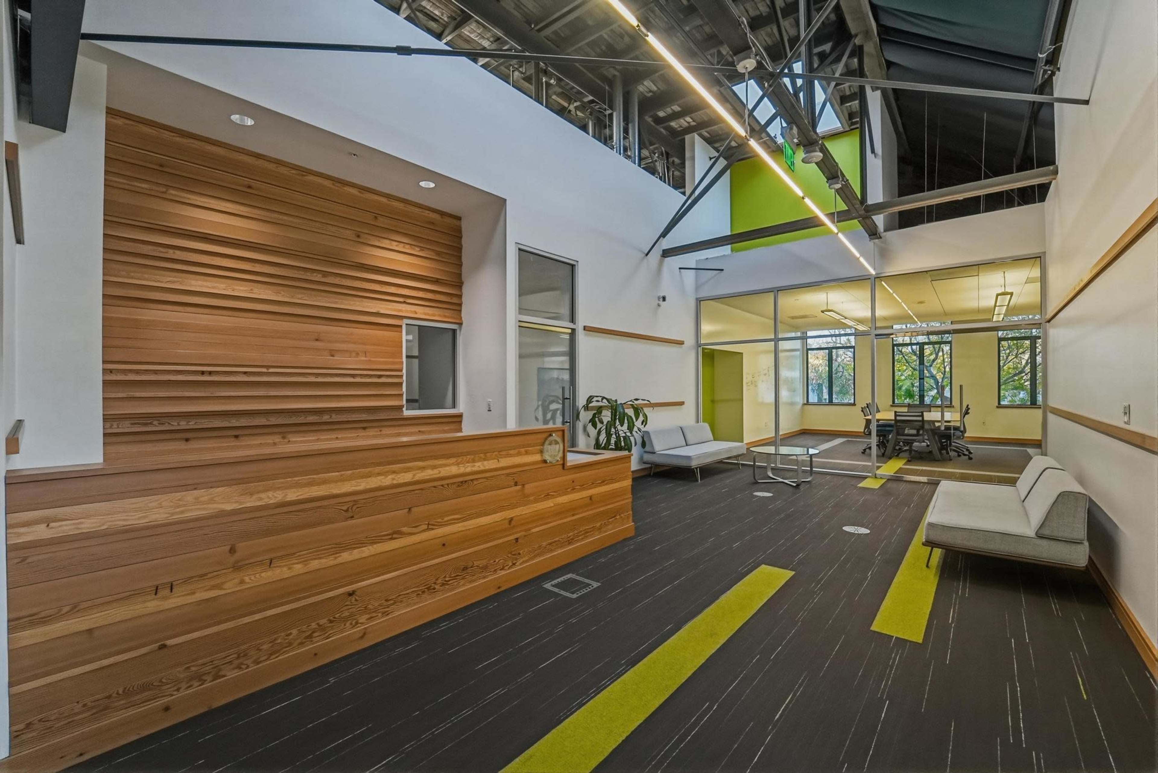 The image shows a modern office interior with wooden paneling, a seating area, and large windows allowing natural light.