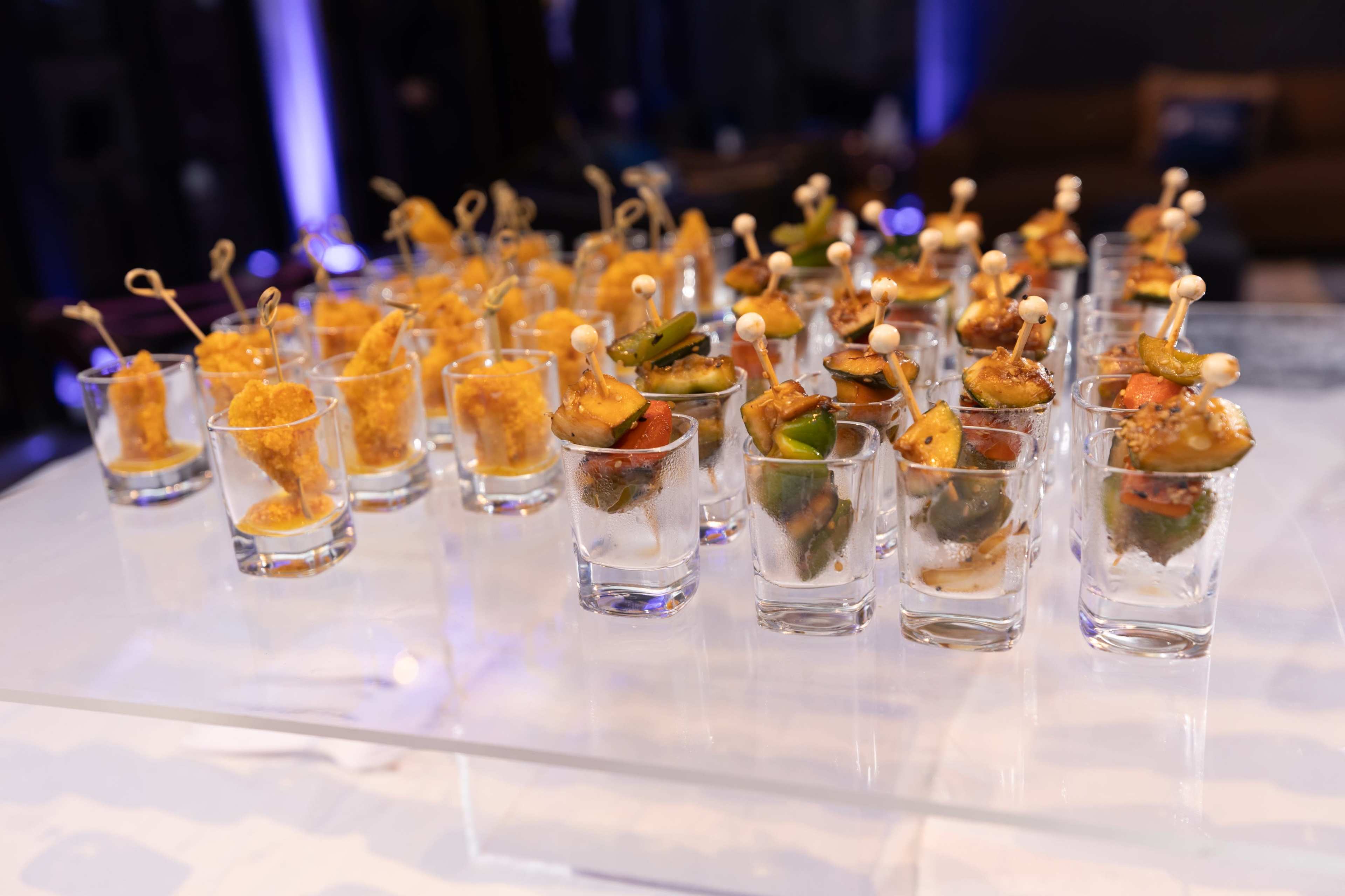 A close-up display of shot glasses filled with assorted appetizers, including skewered vegetables and crispy snacks, arranged on a clear surface.
