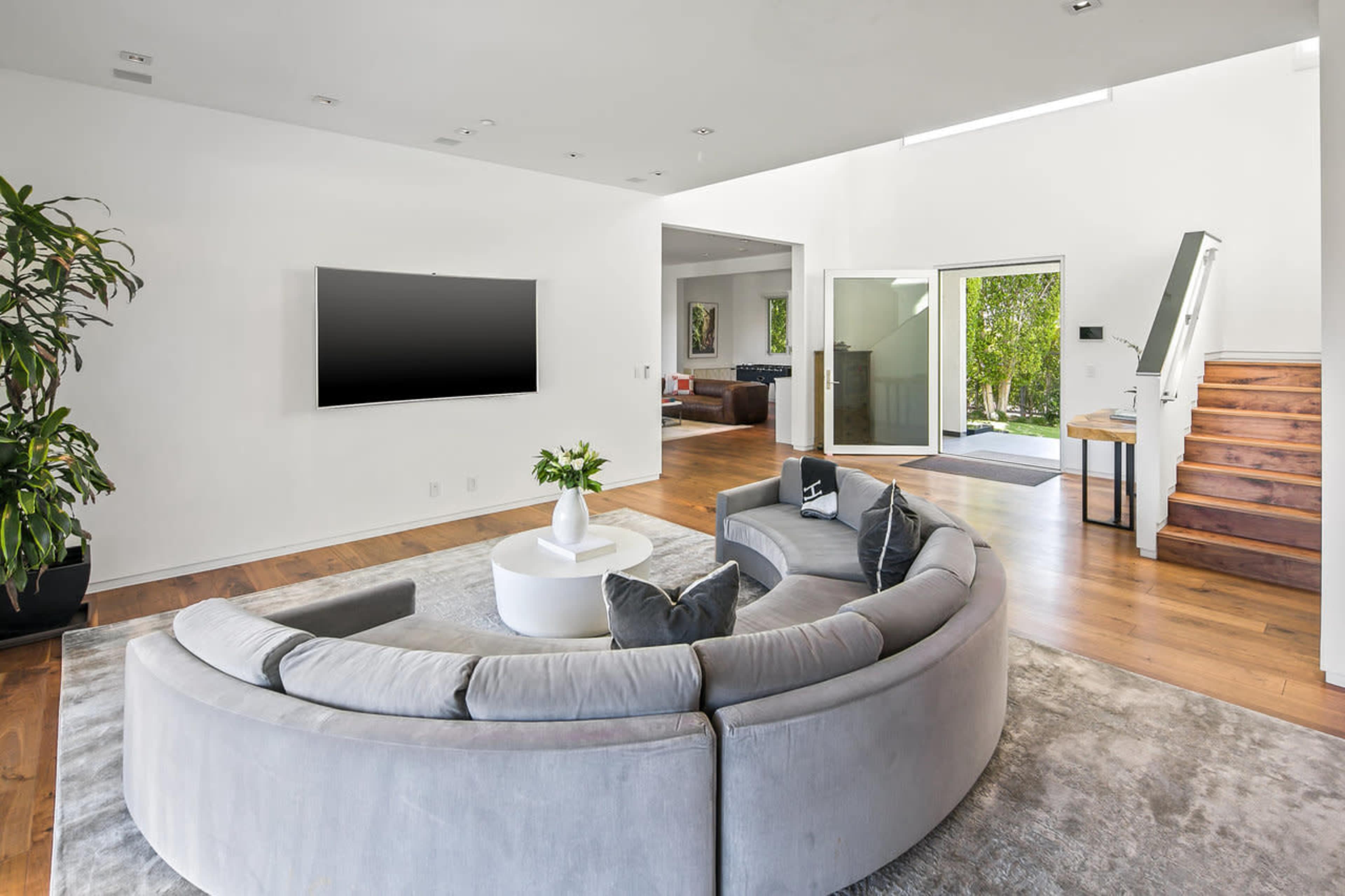 A modern living room features a circular gray couch, a white coffee table, and a wall-mounted TV, with wooden stairs leading to an upper level.