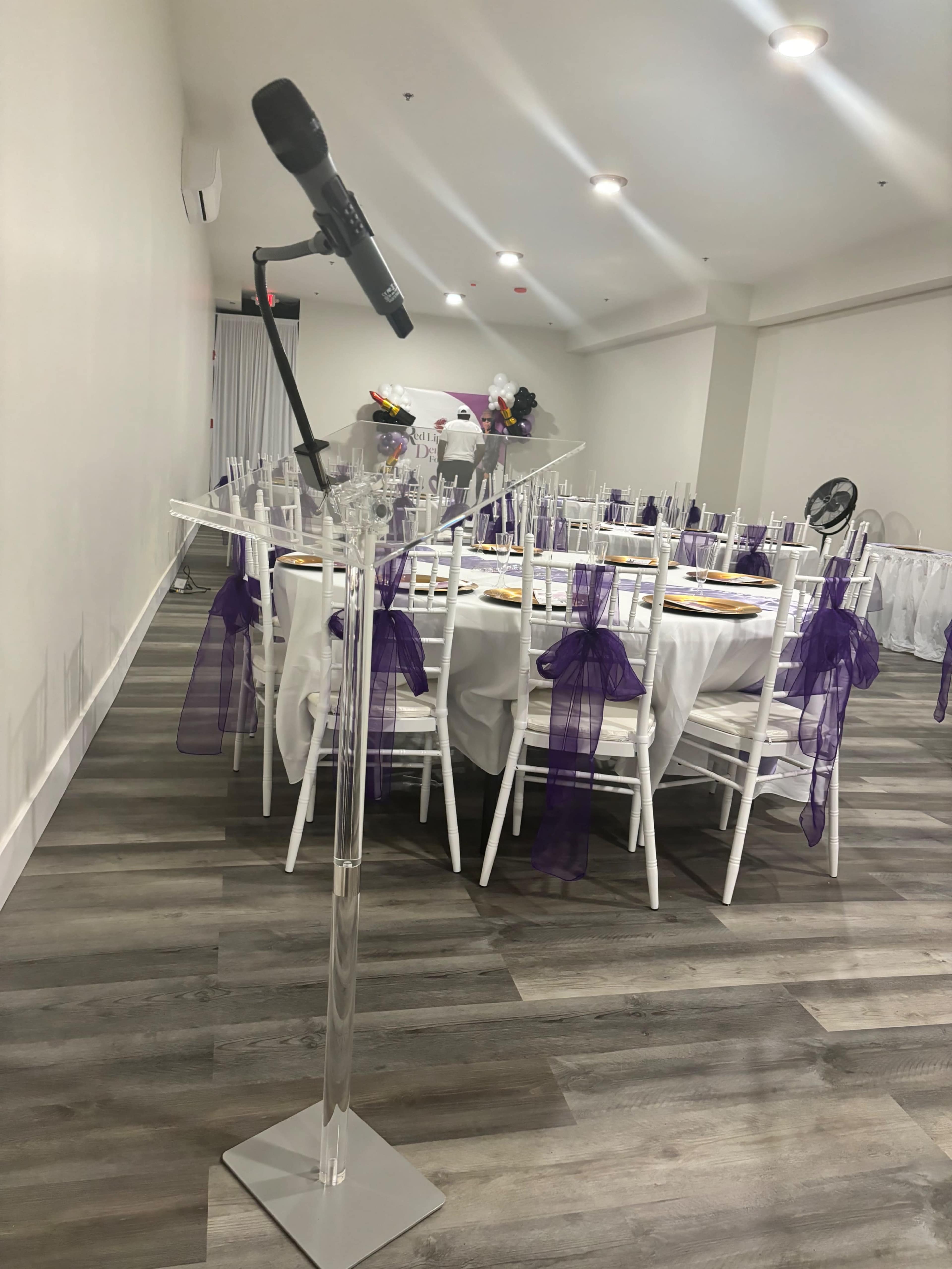 A microphone stands on a clear podium in a banquet room with white tables adorned with purple chairs.