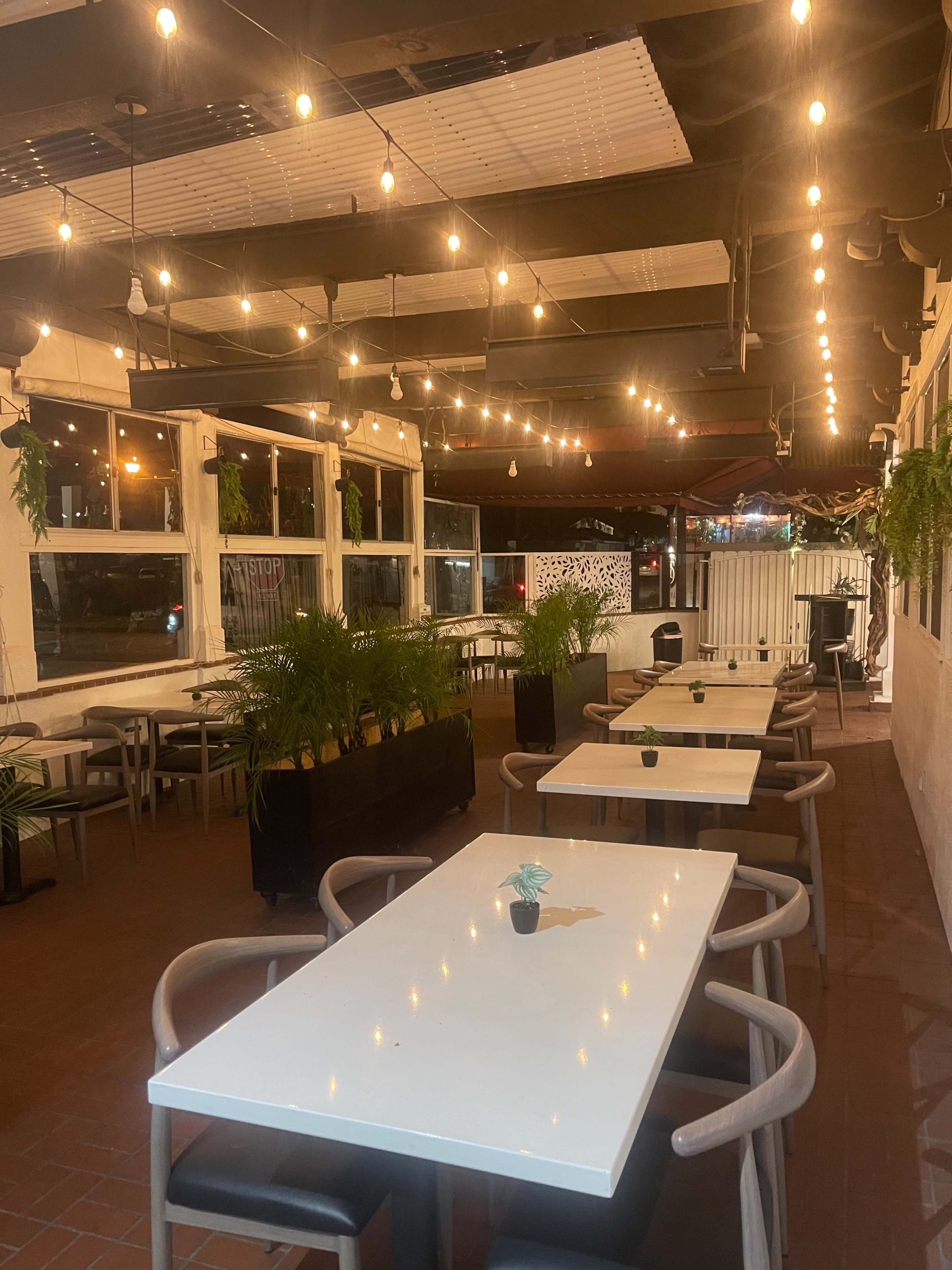 An outdoor restaurant space with several white tables, string lights overhead, and potted plants along the walls.