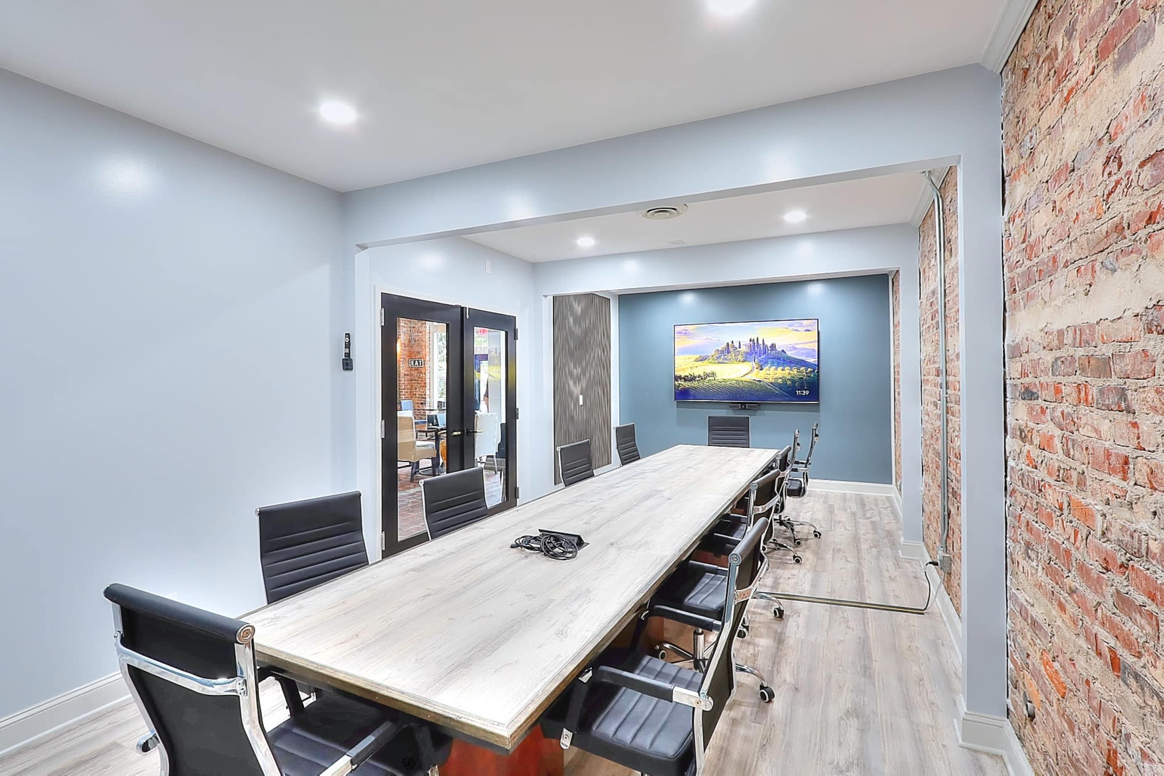 A modern conference room features a long wooden table surrounded by black chairs, with a large screen on one wall and exposed brick accents.