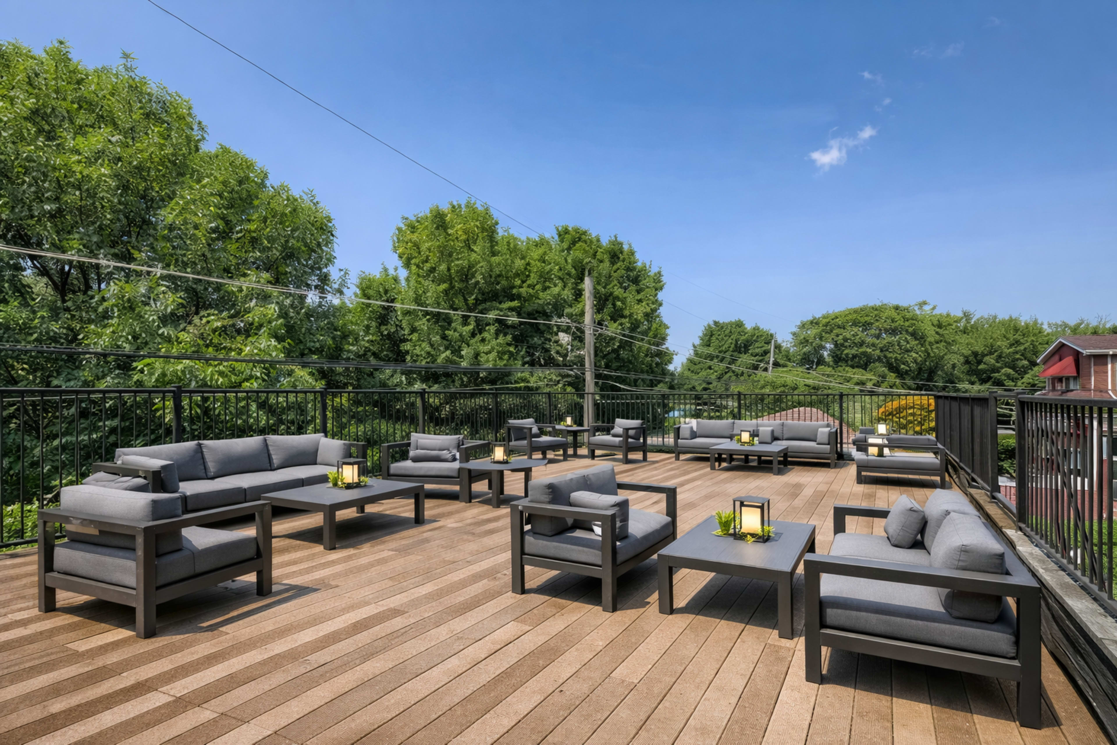 The image shows a rooftop deck furnished with gray couches and tables, surrounded by greenery under a clear blue sky.