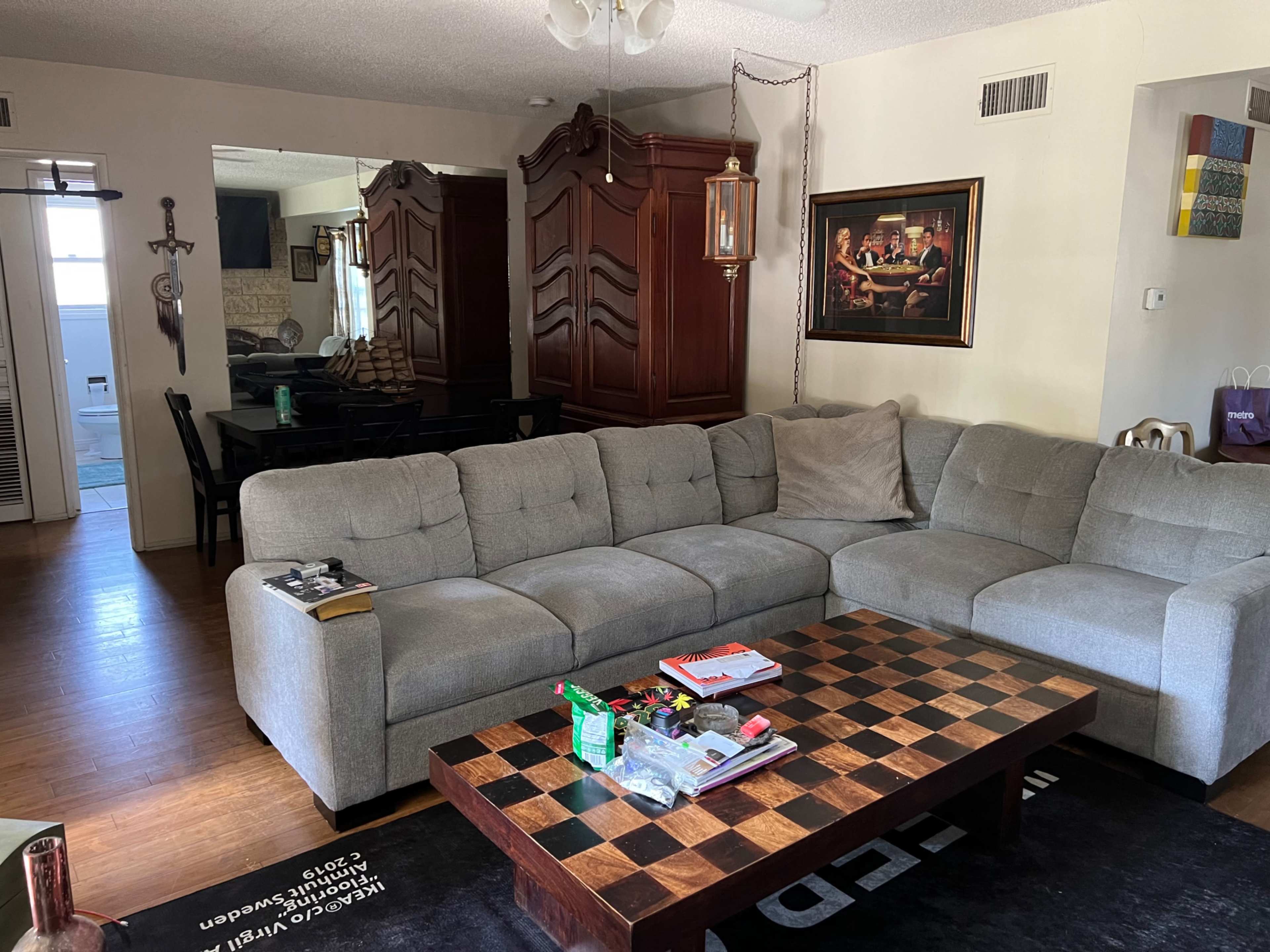 The image shows a living room featuring a gray sectional sofa, a chessboard coffee table, and a dining area with a wooden table in the background.