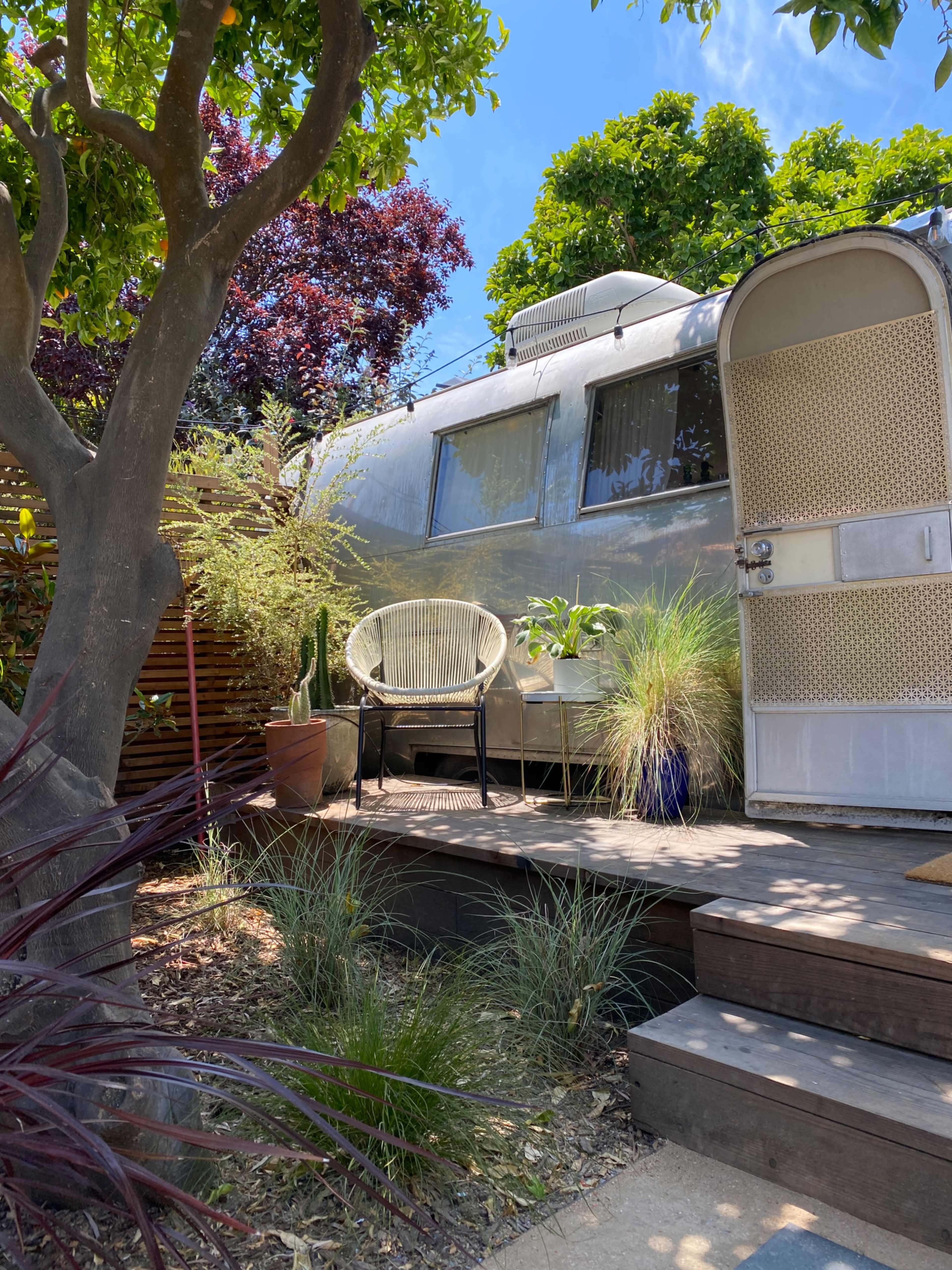 A silver Airstream trailer is parked beside a wooden deck surrounded by plants and trees.