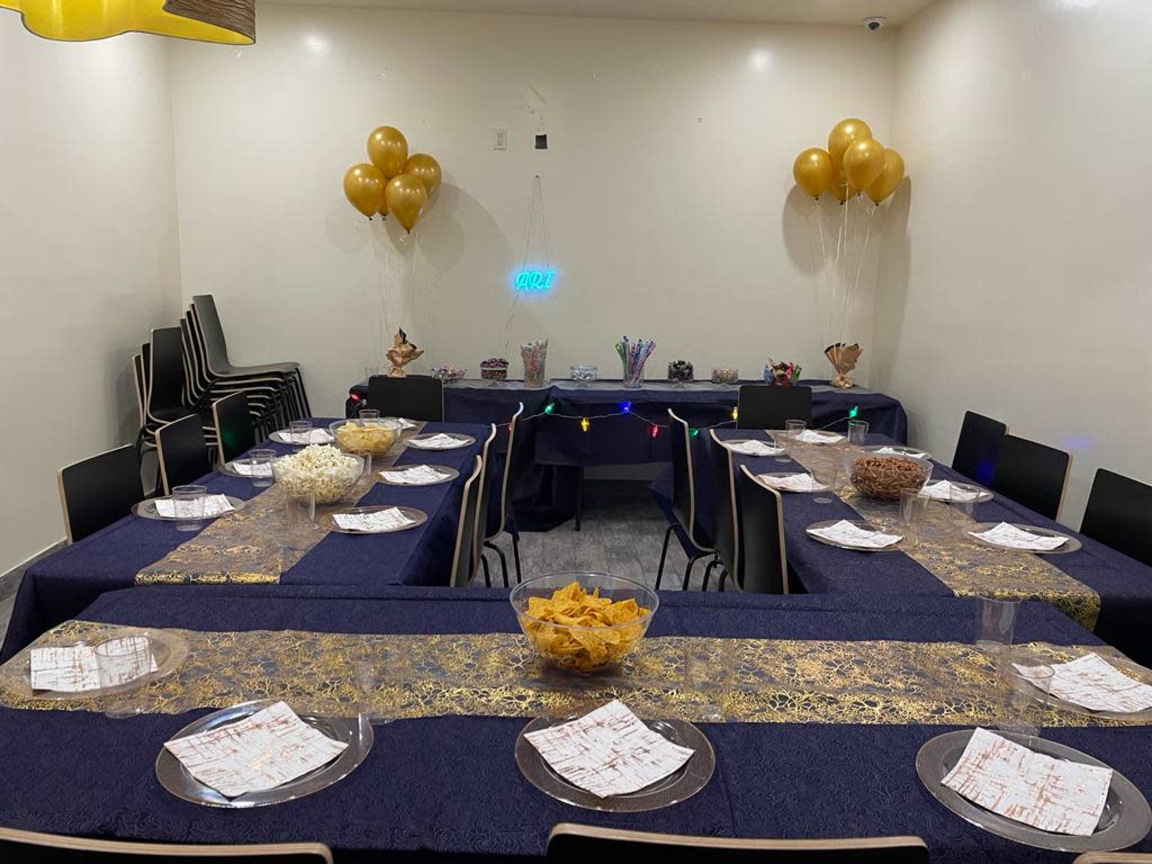 A banquet room is set up with several tables covered in blue and gold linens, featuring an assortment of snacks and decorations, including balloons and colorful lights.