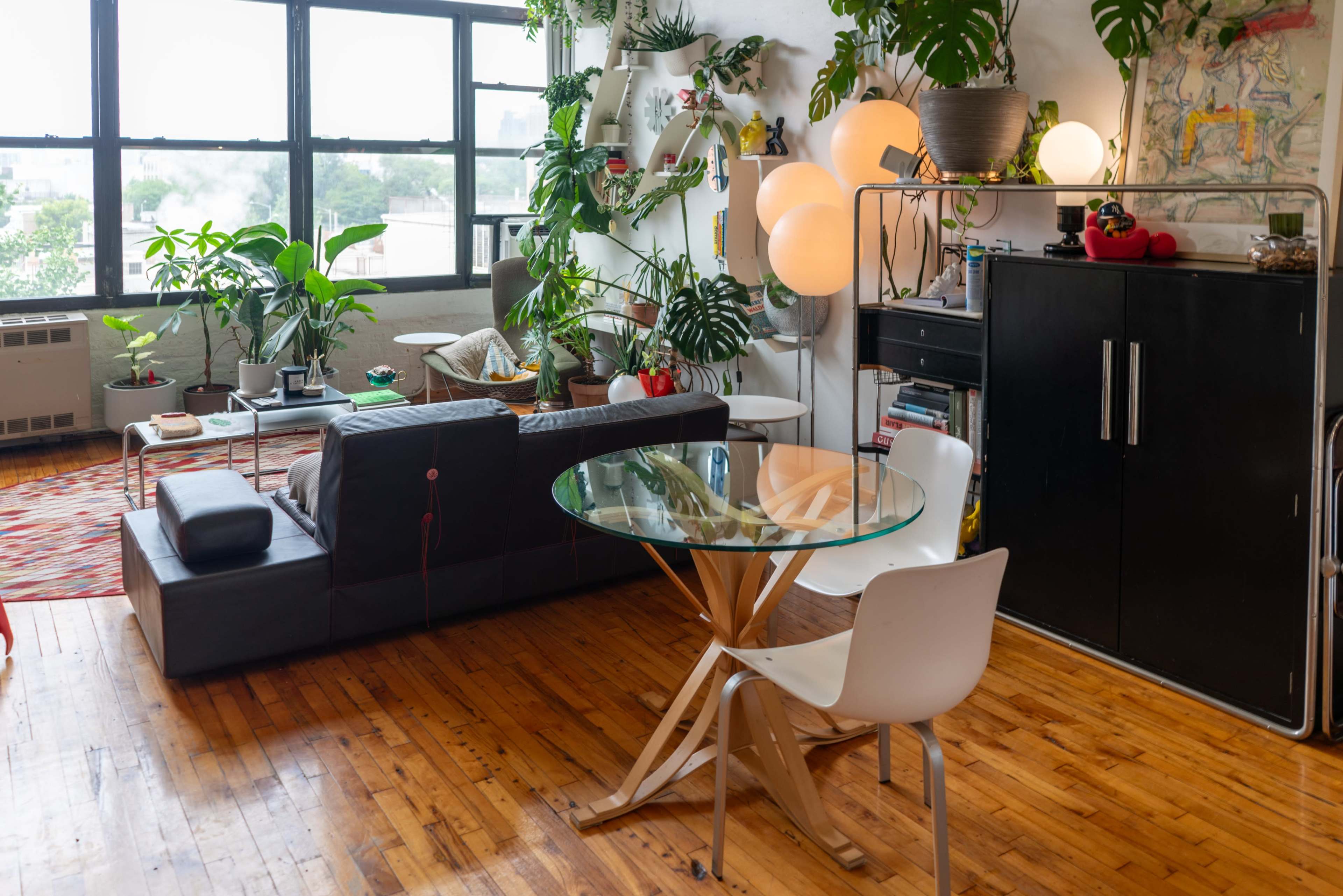 The image shows a modern living space featuring a black sofa, a glass dining table with wooden legs, potted plants, and a black storage unit against the wall.