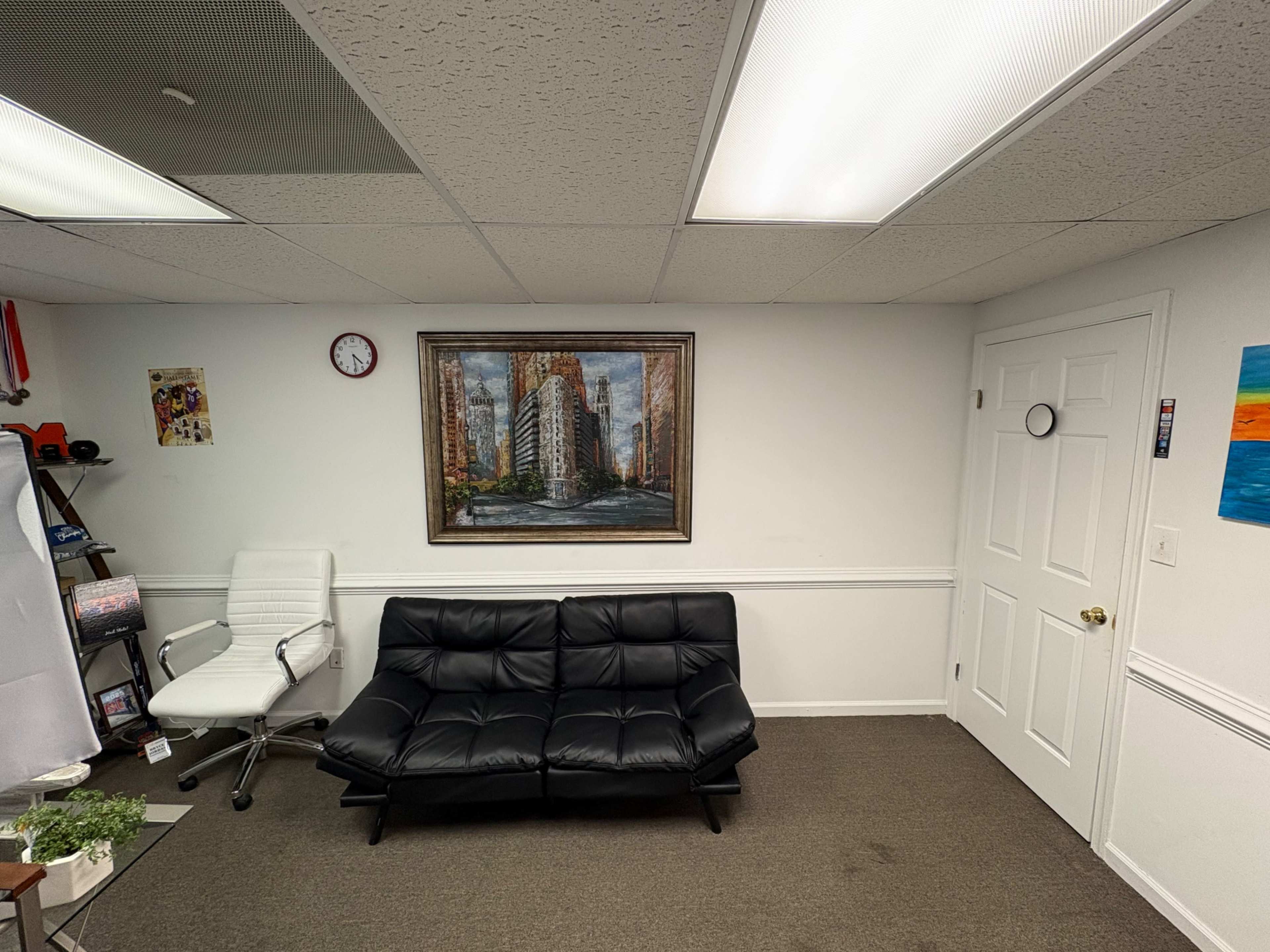 The image shows a small office space featuring a black sofa, a white chair, a wall clock, a framed painting, and a closed door.