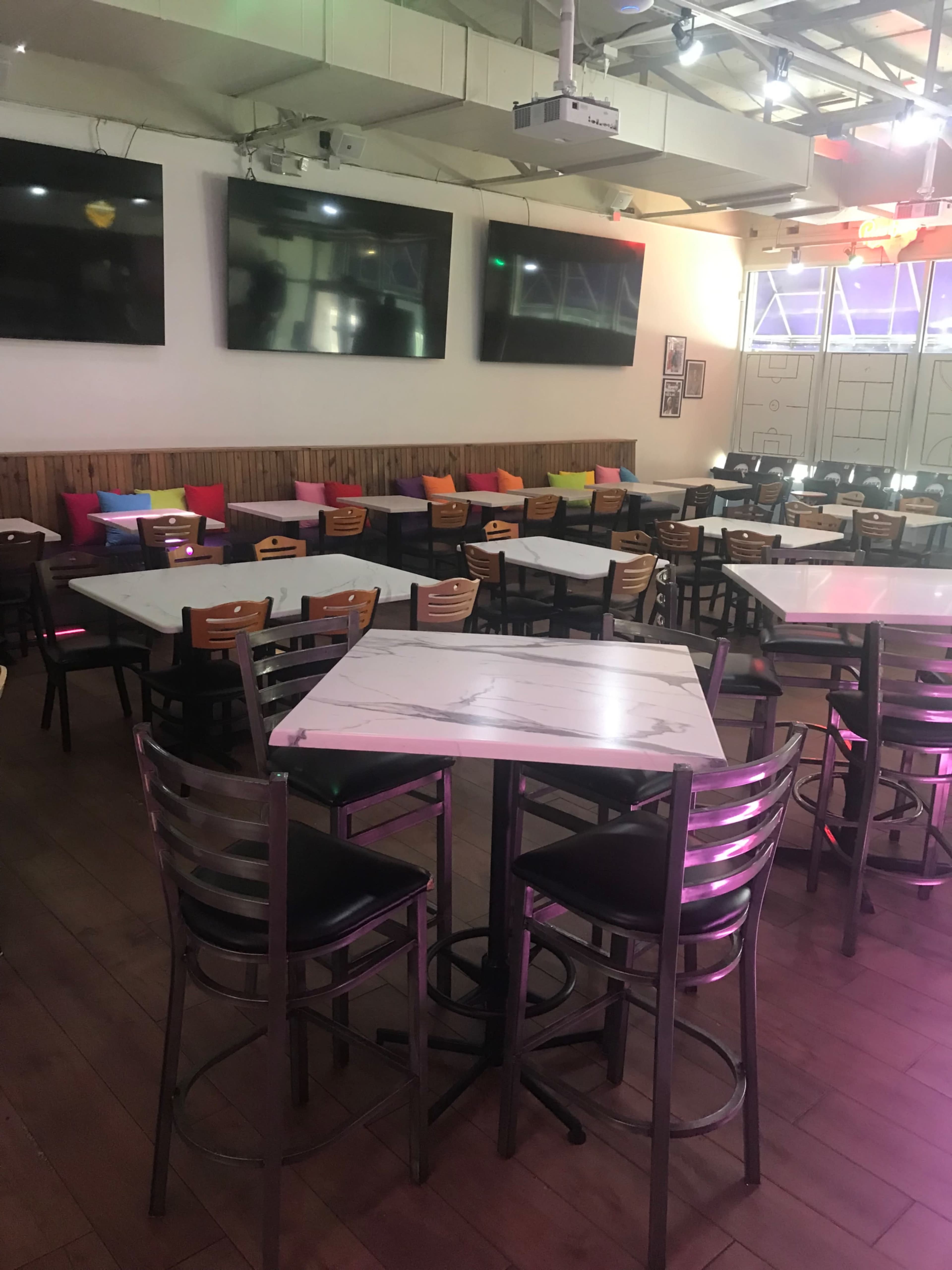 The image shows a spacious restaurant interior with several tables and chairs arranged around, featuring colorful cushions along the back wall and multiple large screens mounted above.