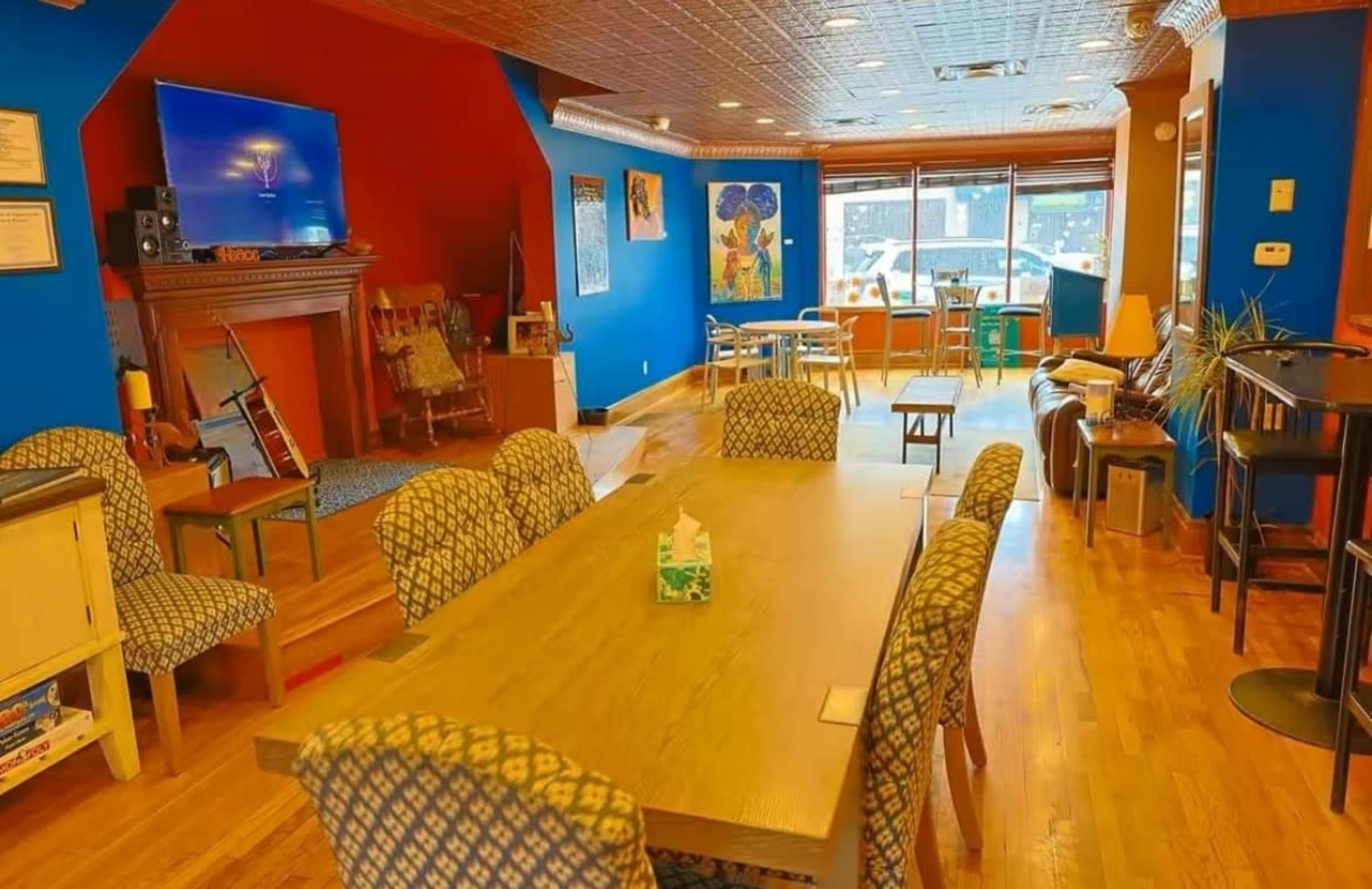 The image shows the interior of a cozy café with a large wooden table, colorful walls, and a variety of seating arrangements.