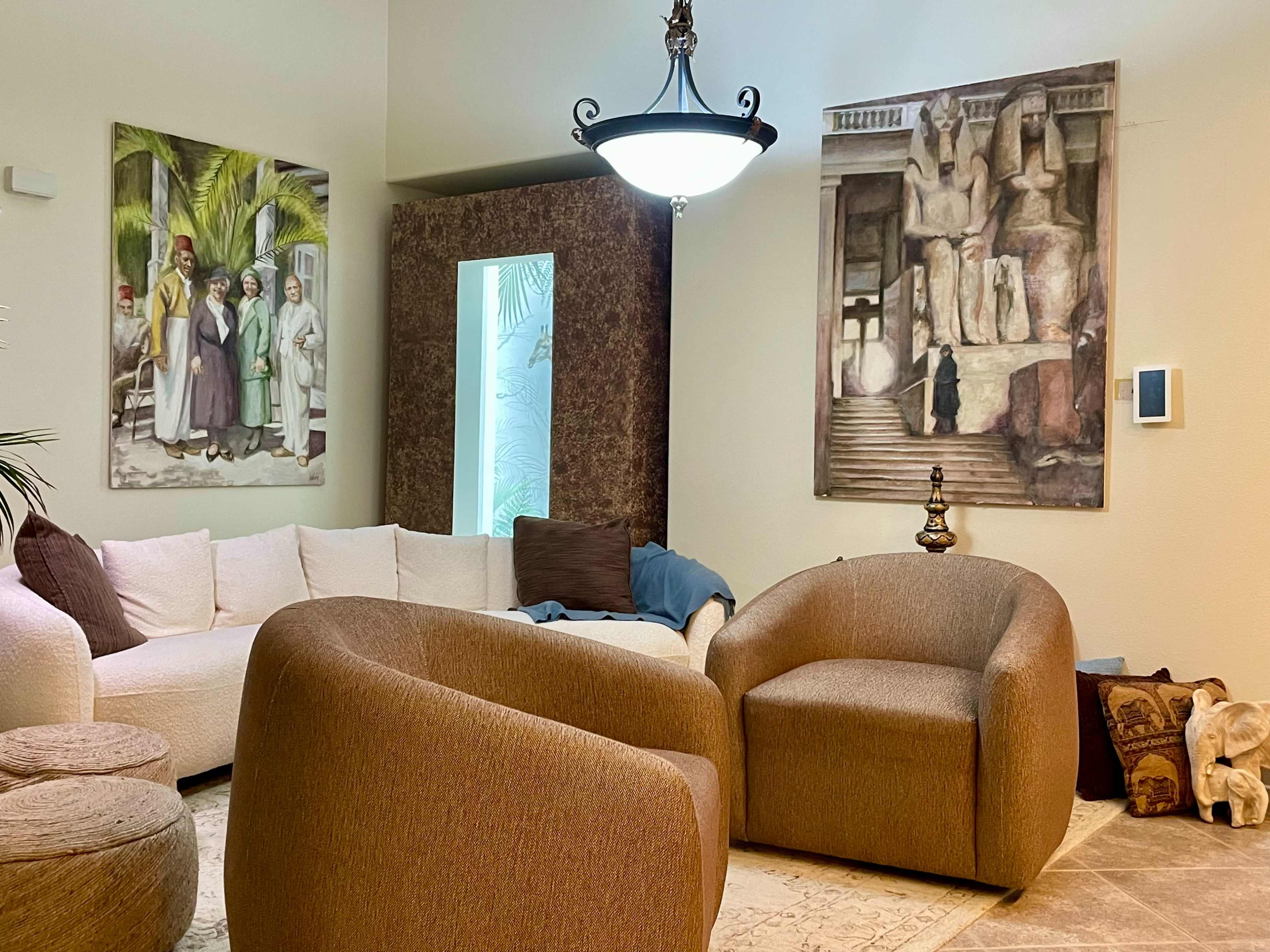 The image shows a living room with two curved brown chairs, a light-colored sofa, and two large framed paintings on the walls, along with a decorative lamp and a small table.