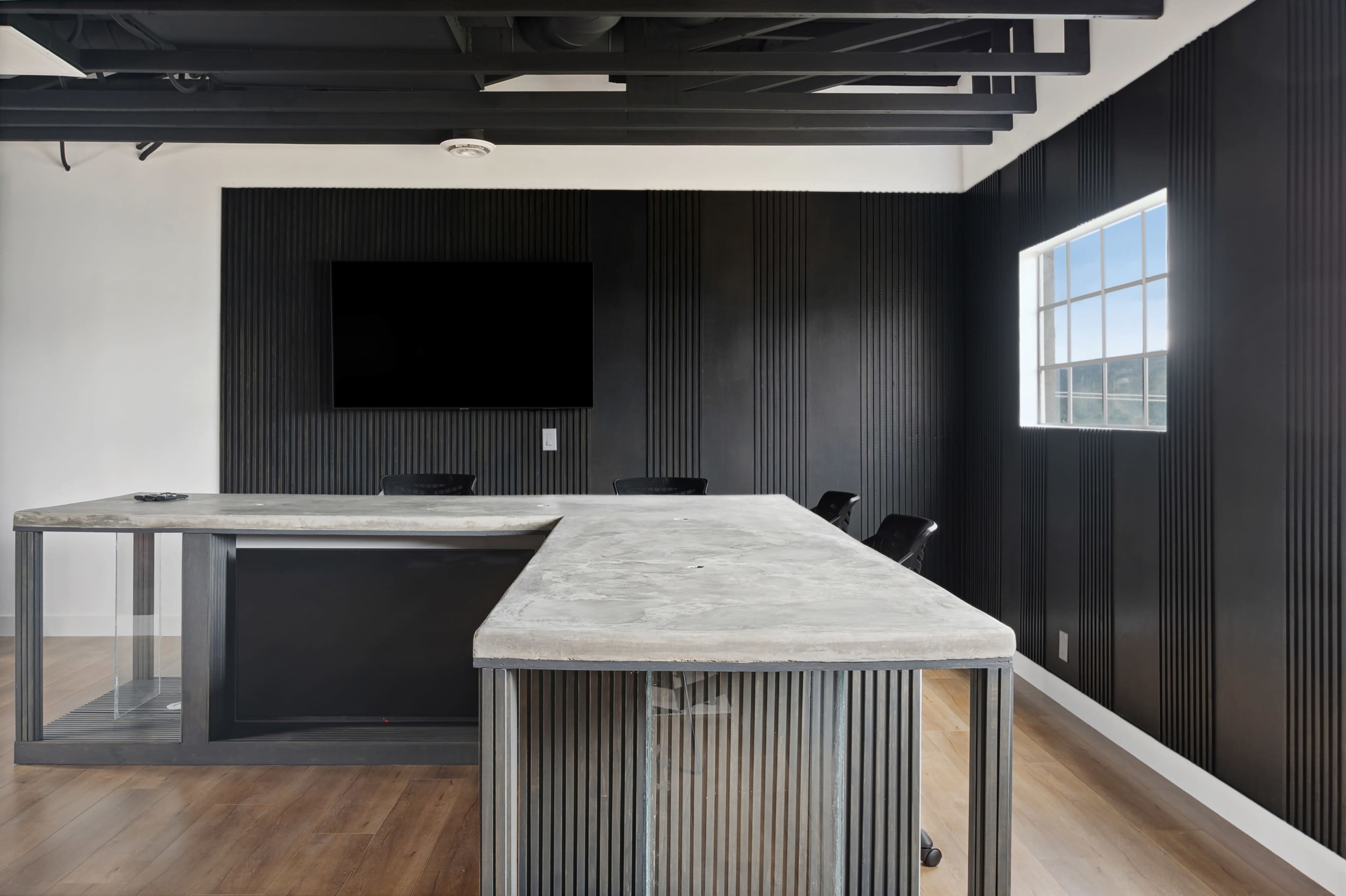 The image shows a modern office space featuring a concrete bar-style counter, black paneled walls, a large wall-mounted screen, and a window with natural light.