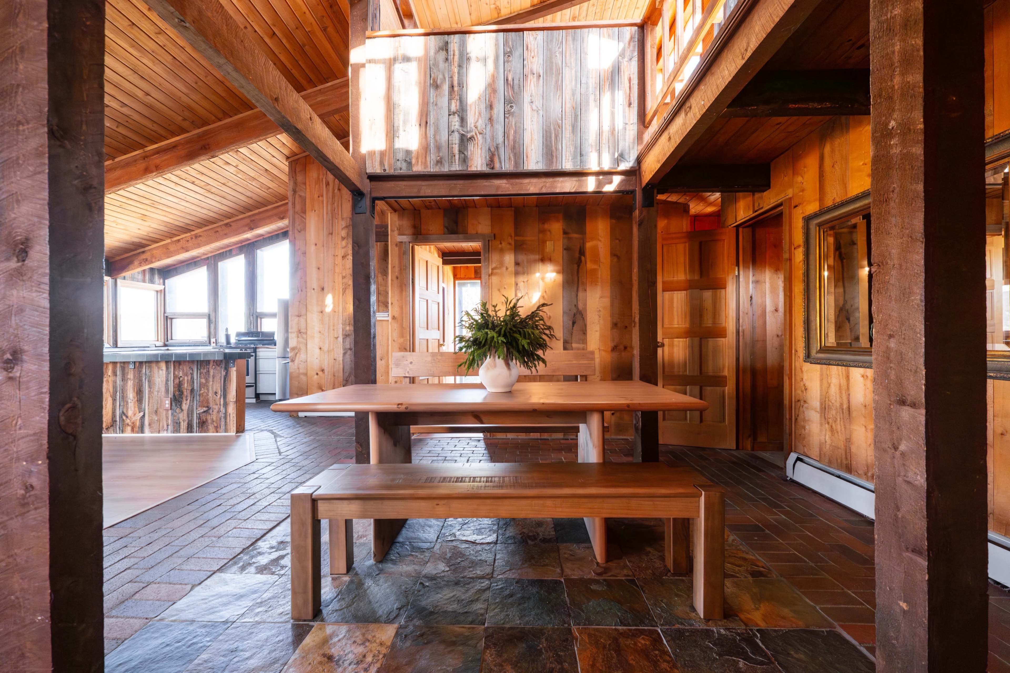 The image depicts a rustic dining area with a wooden table and bench, surrounded by walls of natural wood and stone flooring, illuminated by large windows above.