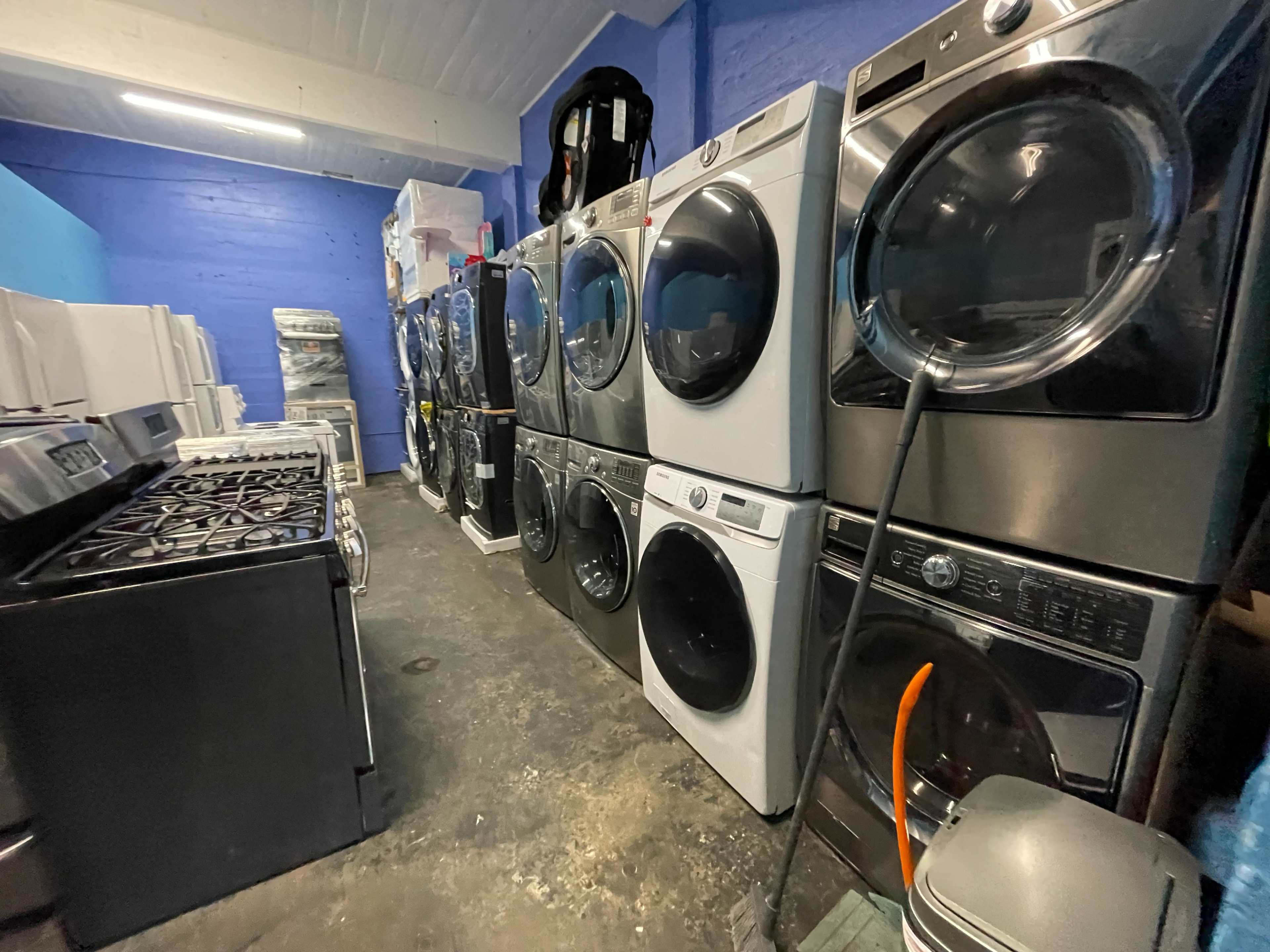 A row of various washing machines and dryers is displayed in a warehouse, alongside a black gas stove.