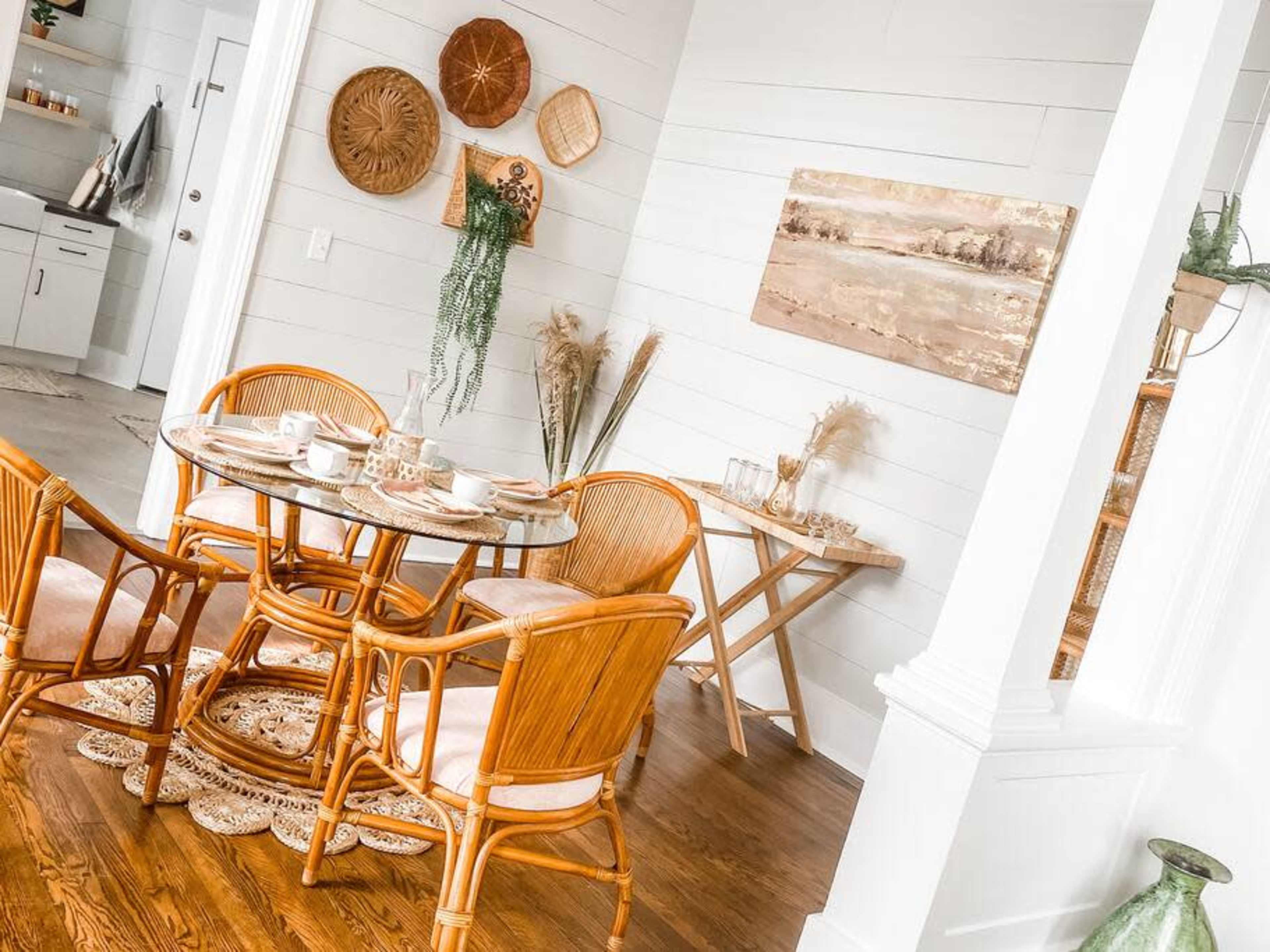 A dining area features a round table surrounded by four rattan chairs, decorated with plates and cups, alongside a wall adorned with woven art and a landscape painting.