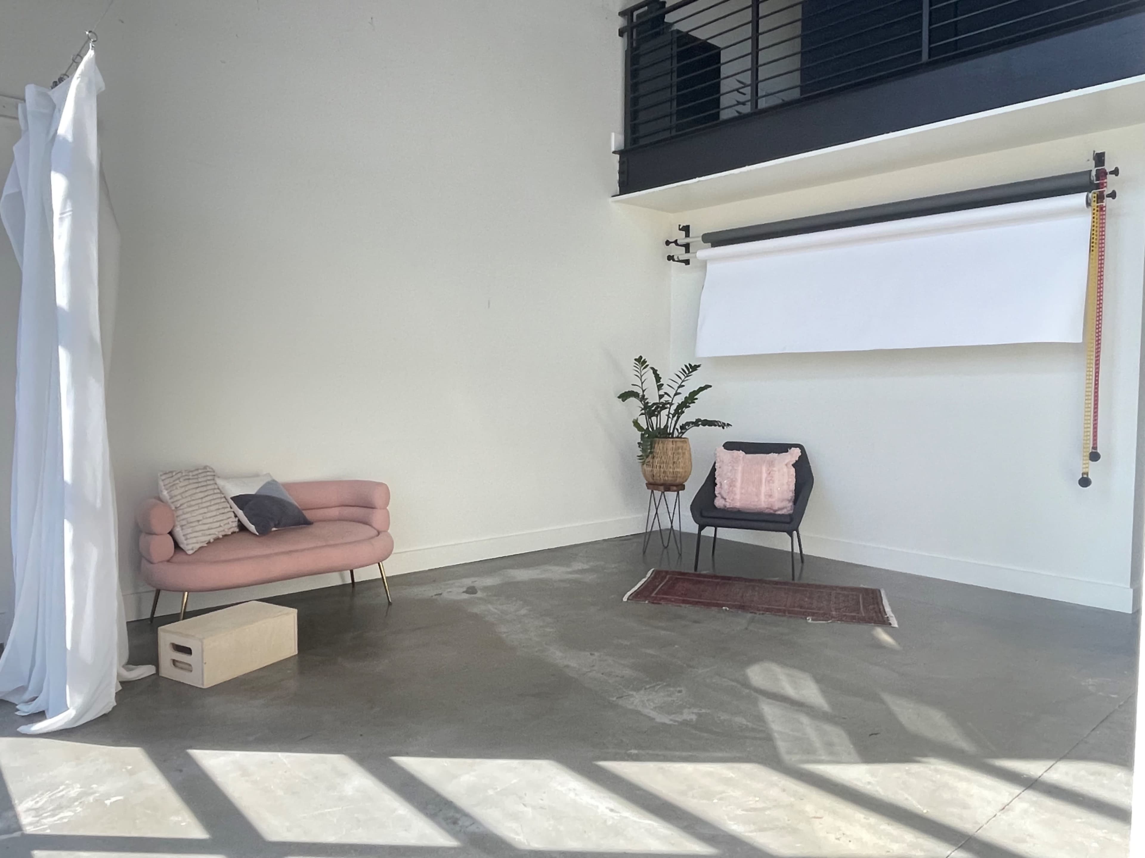 A spacious room features a pink sofa, a black chair with a pillow, a small box, a potted plant, and a white backdrop, all set against a neutral wall and floor.