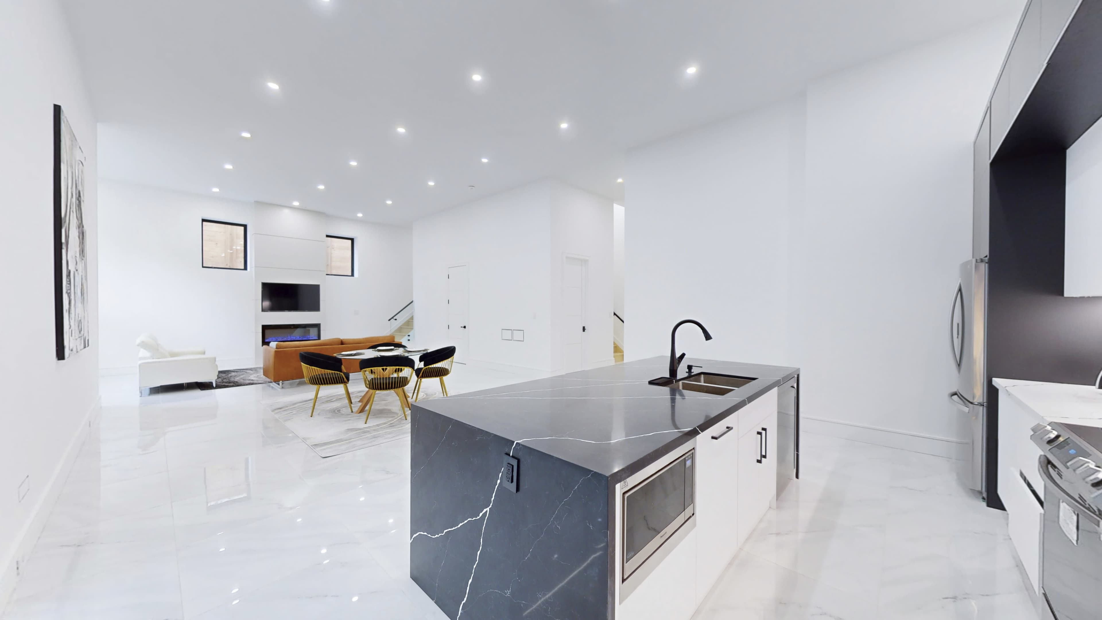 A modern, open-concept kitchen and living area with a large island, sleek appliances, and a bright, airy atmosphere created by white walls and marble flooring.