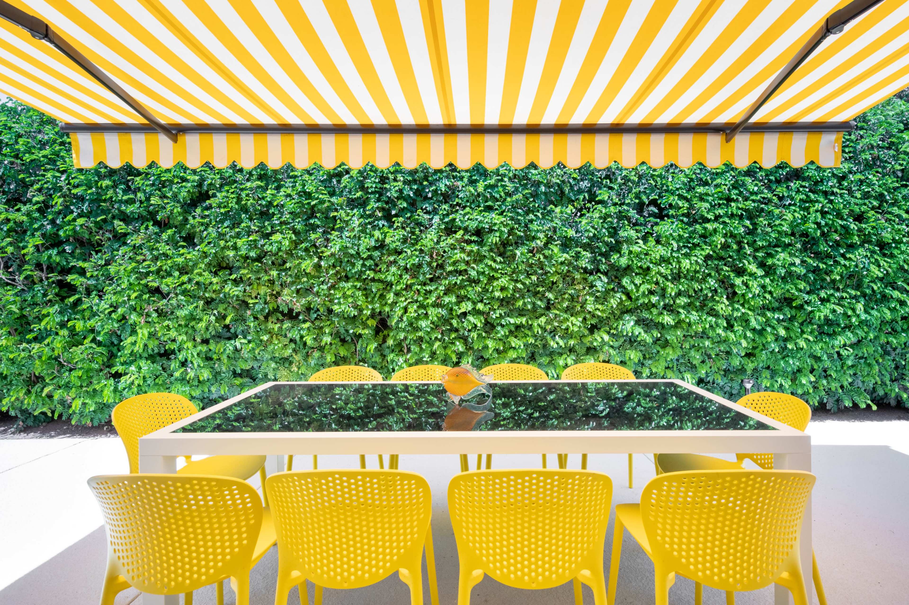 A dining table with a glass top and yellow chairs is positioned under a striped awning, surrounded by lush green foliage.