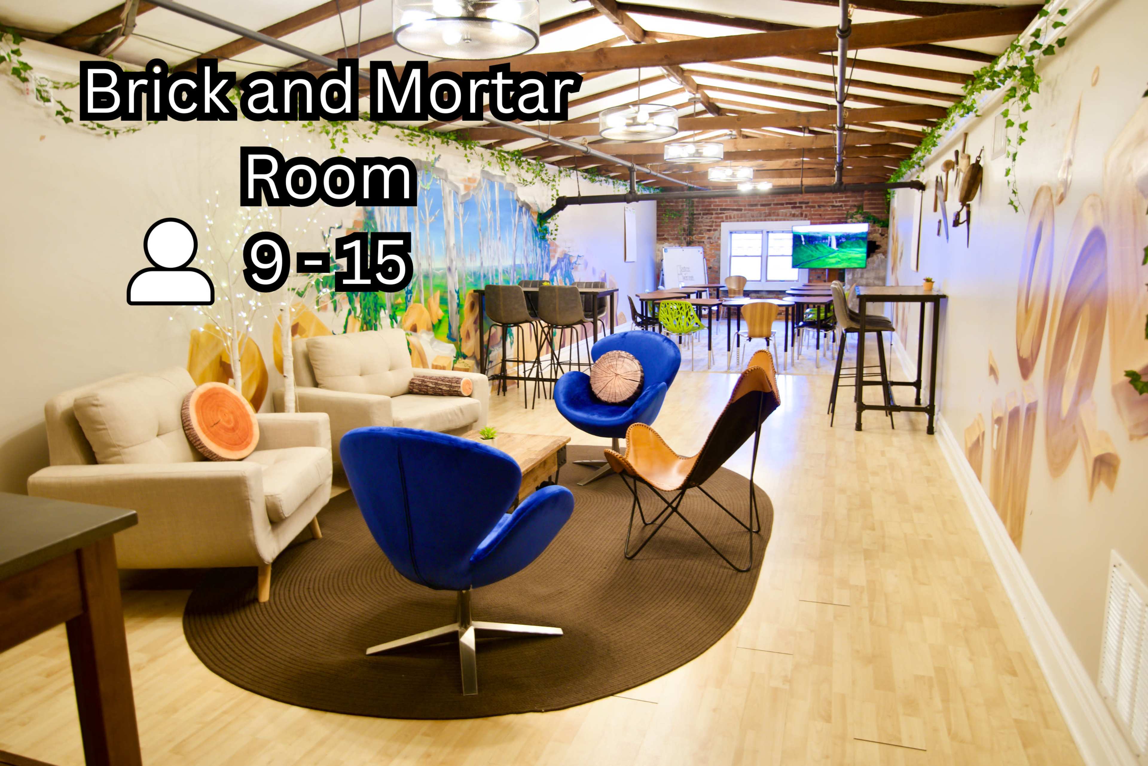 The image shows a spacious room with seating areas, including chairs and sofas, colorful wall art, and a workspace with tables and a television.