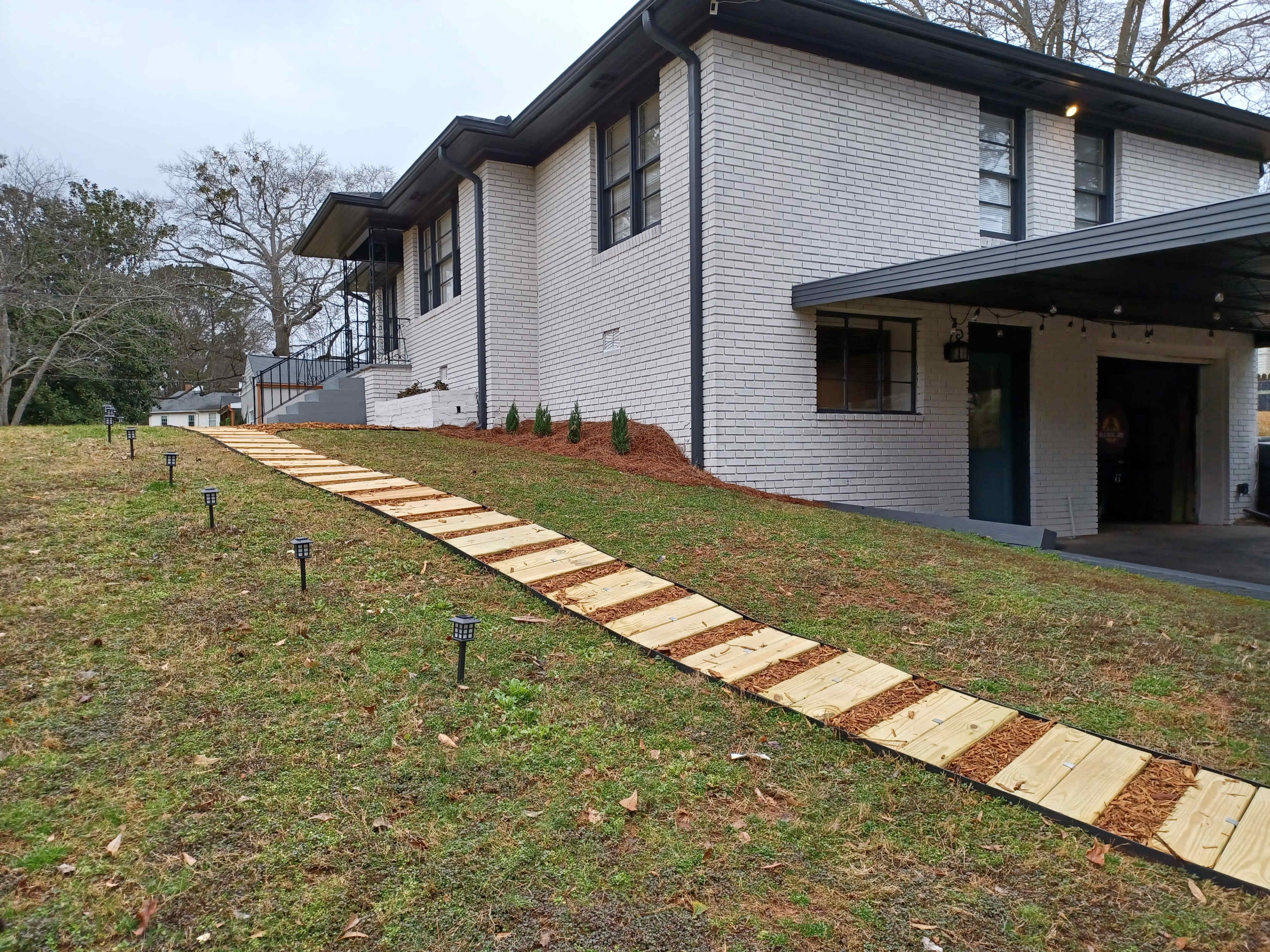 The image shows a wooden pathway leading up a grassy slope to a modern, two-story house with a white brick exterior.