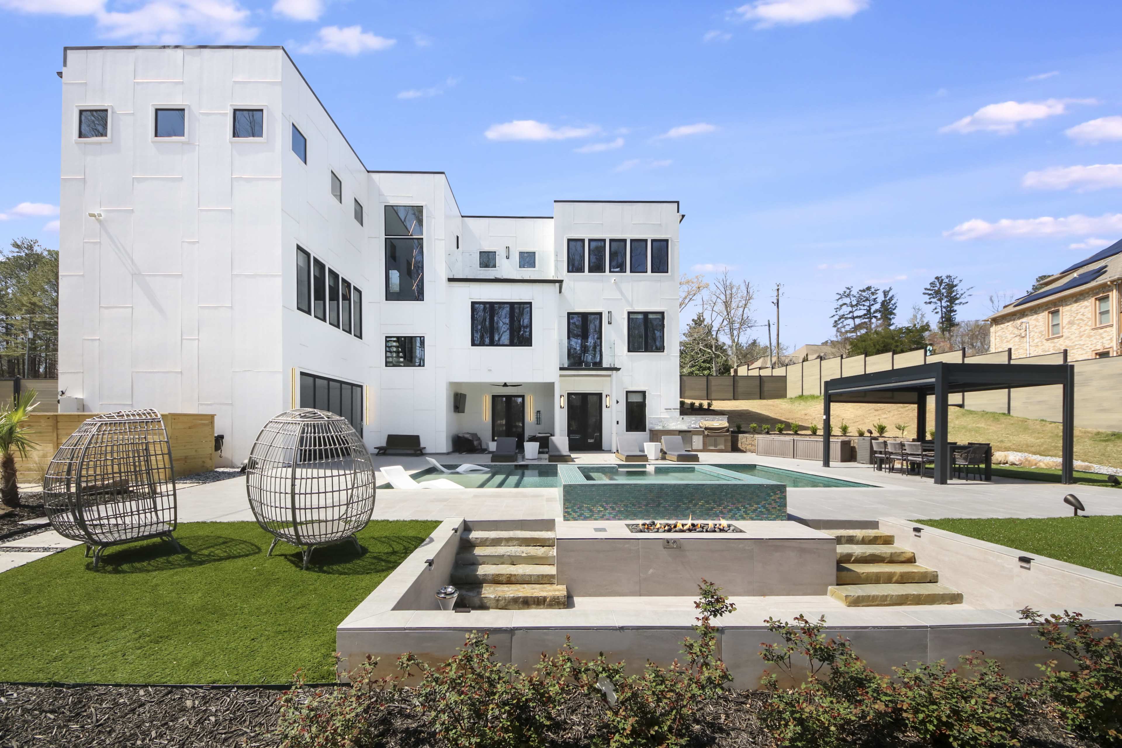 A modern two-story house with large windows, surrounded by a patio, a landscaped yard, and a pool area, featuring two woven chairs and a fire pit.