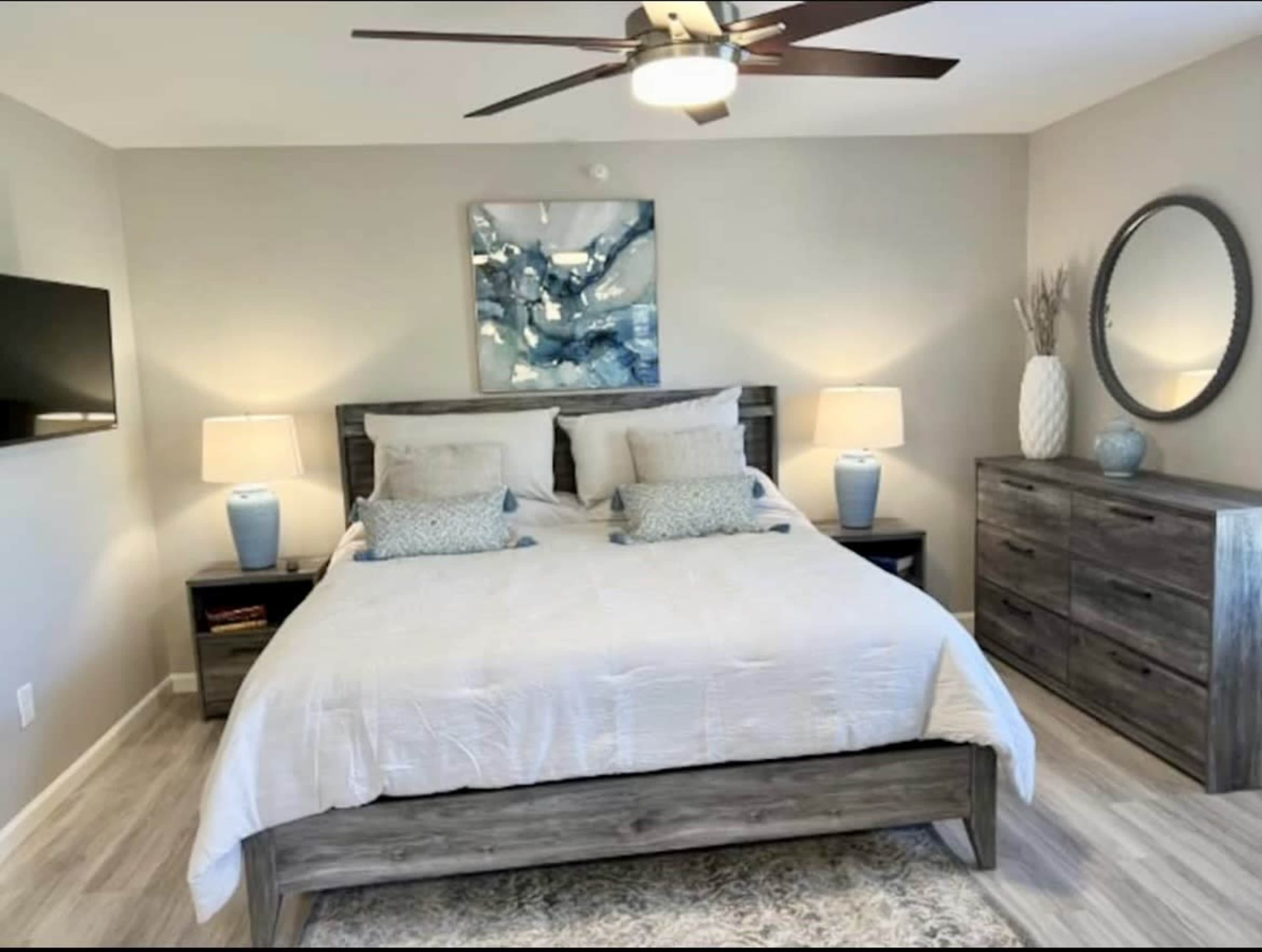 A neatly arranged bedroom featuring a queen-sized bed, two bedside tables with lamps, a dresser, and a wall-mounted television.