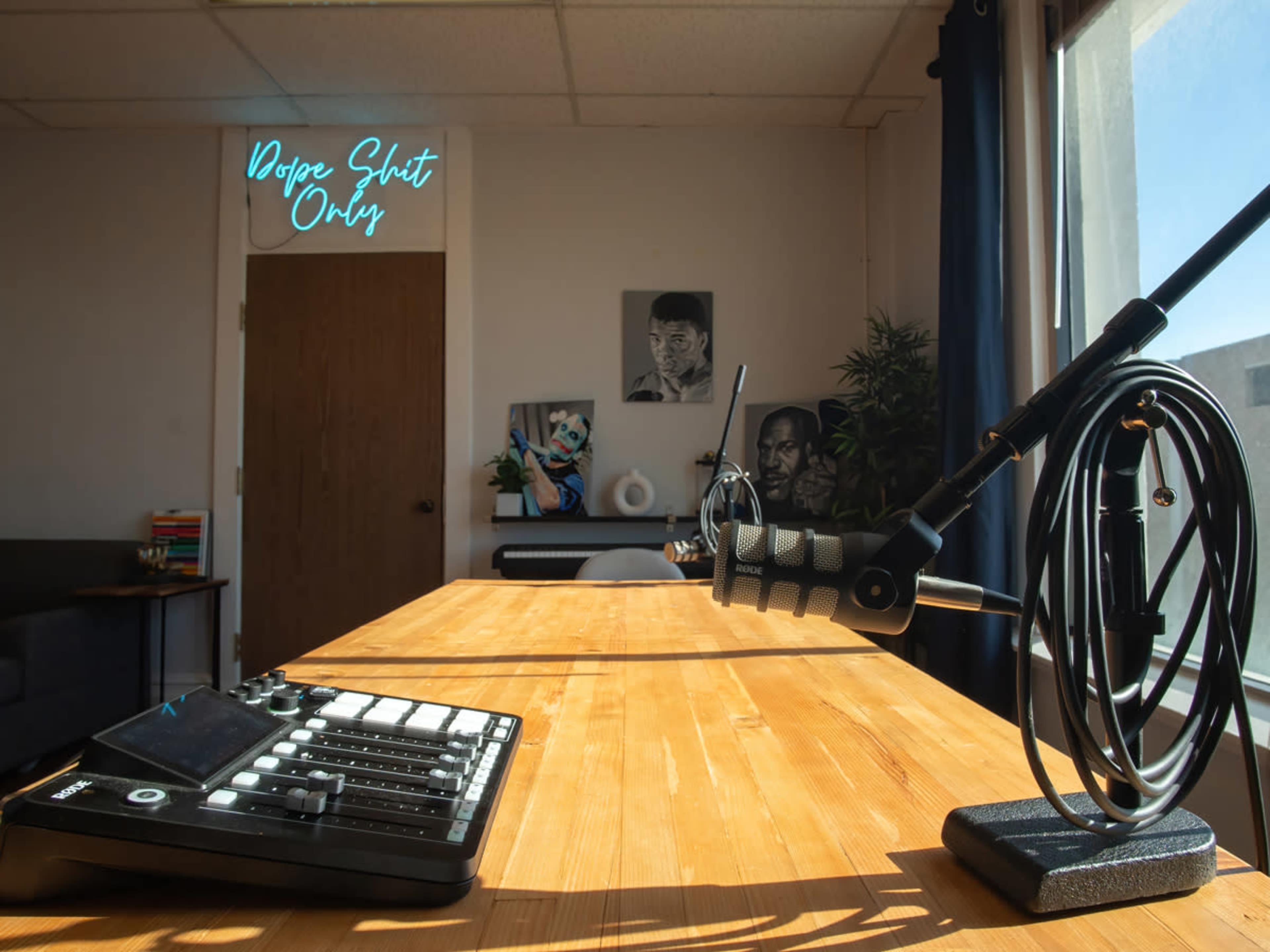 A well-lit room featuring a wooden table with a microphone on a stand and a control panel, alongside a wall with framed artwork and a neon sign that reads "Dodge Shunt Only."