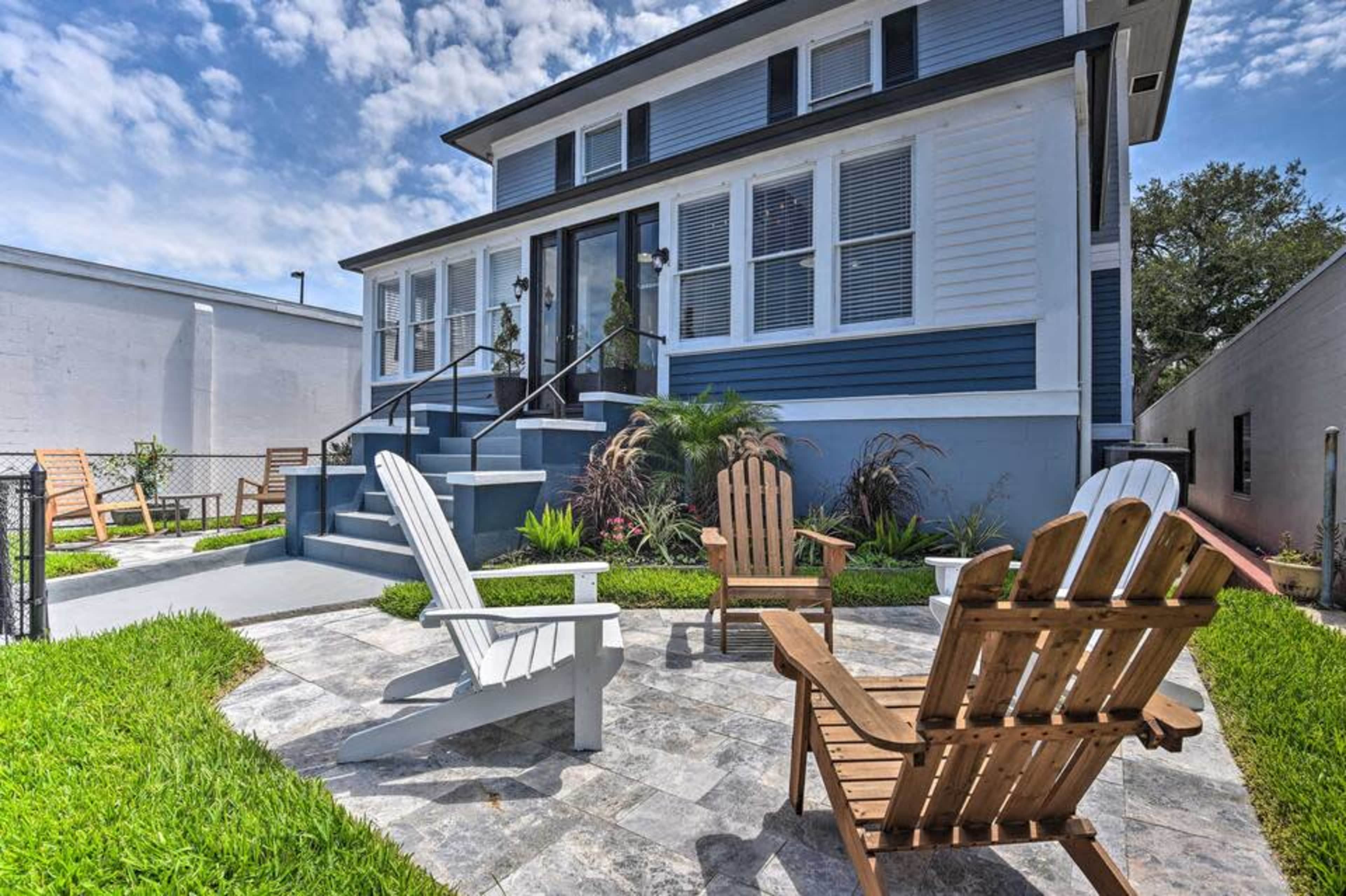 A blue and white house features a front porch with steps leading to a paved seating area surrounded by Adirondack chairs and landscaped greenery.