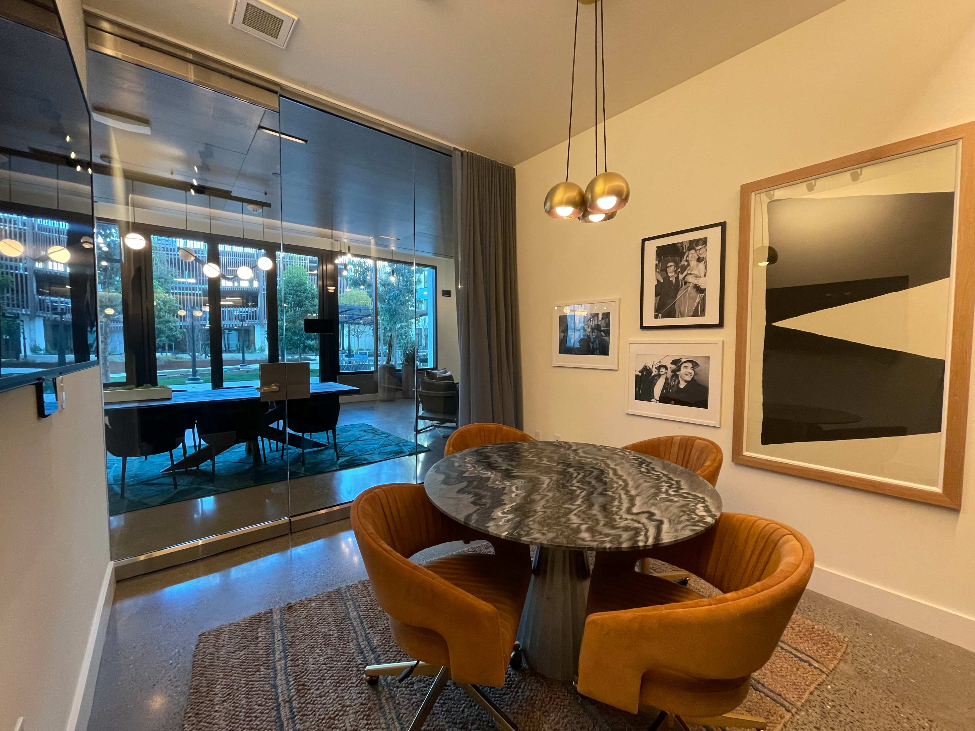 A dining area features a circular marble table surrounded by orange chairs, with a wall adorned by framed artwork, and a glass wall revealing an outdoor view.