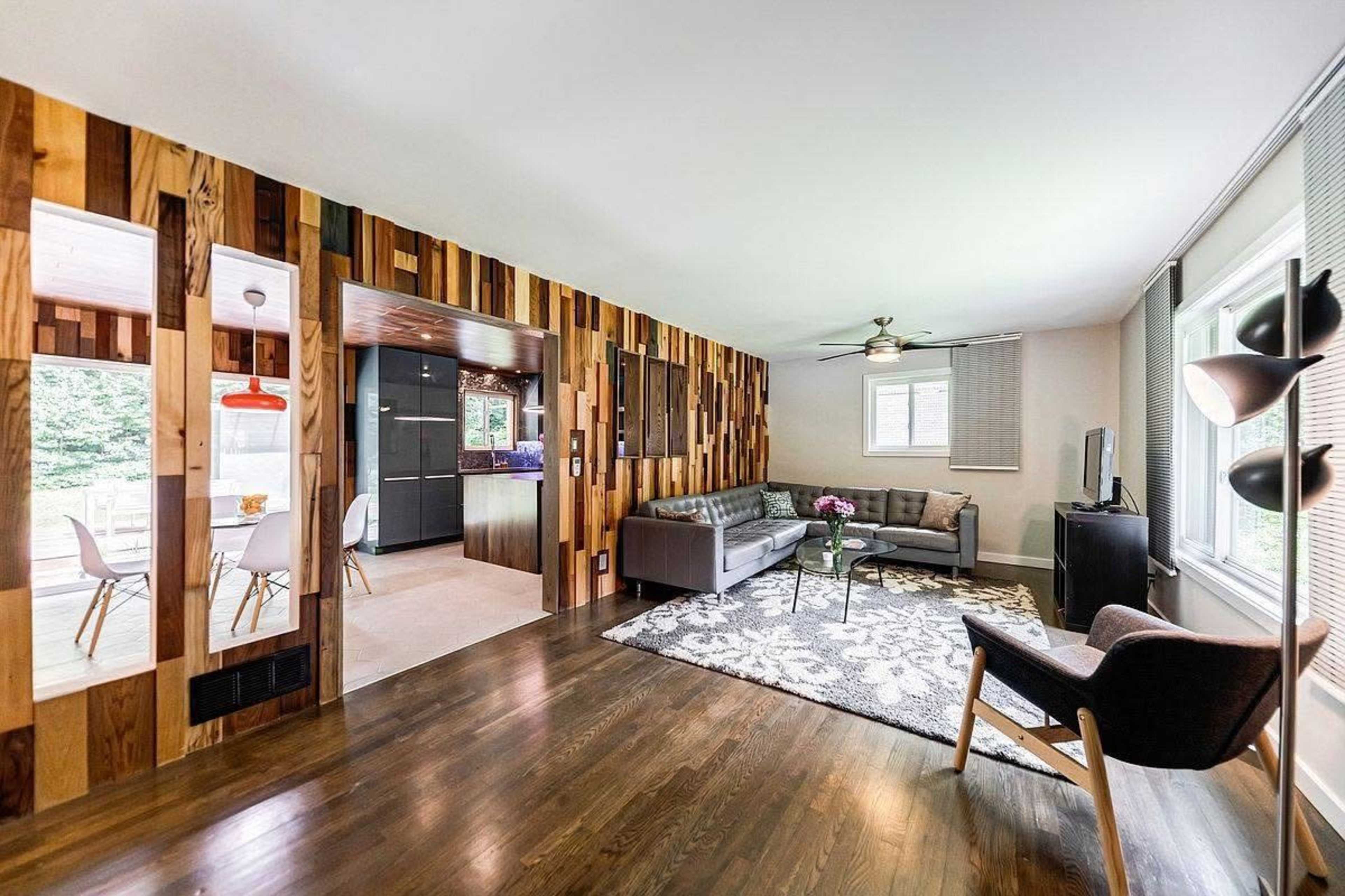 The image shows a modern living room with wooden accent walls, a gray sectional sofa, and a patterned rug, leading into a kitchen area visible through an open space.