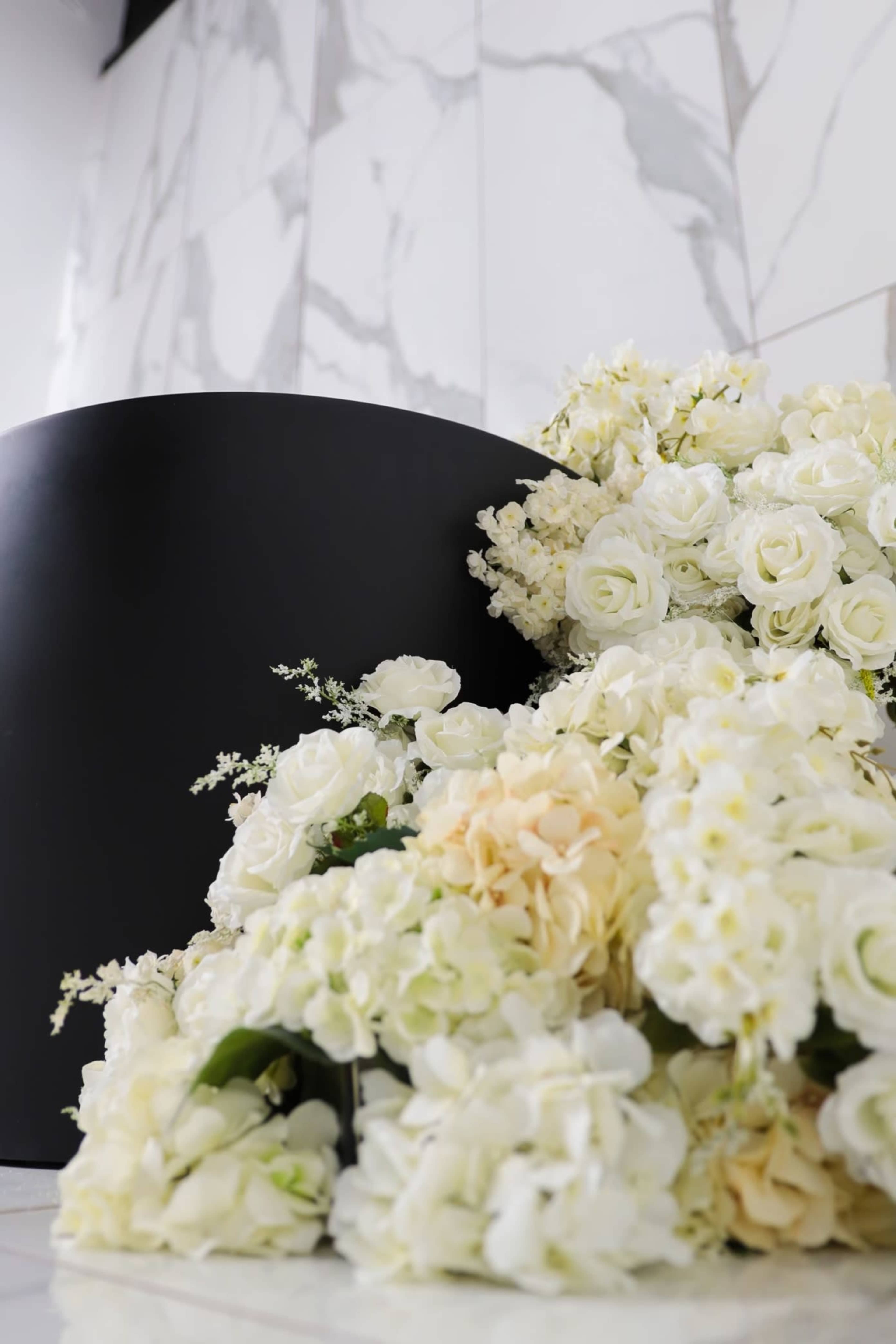 A large arrangement of white flowers, including roses and hydrangeas, is positioned next to a sleek black display stand against a marble background.