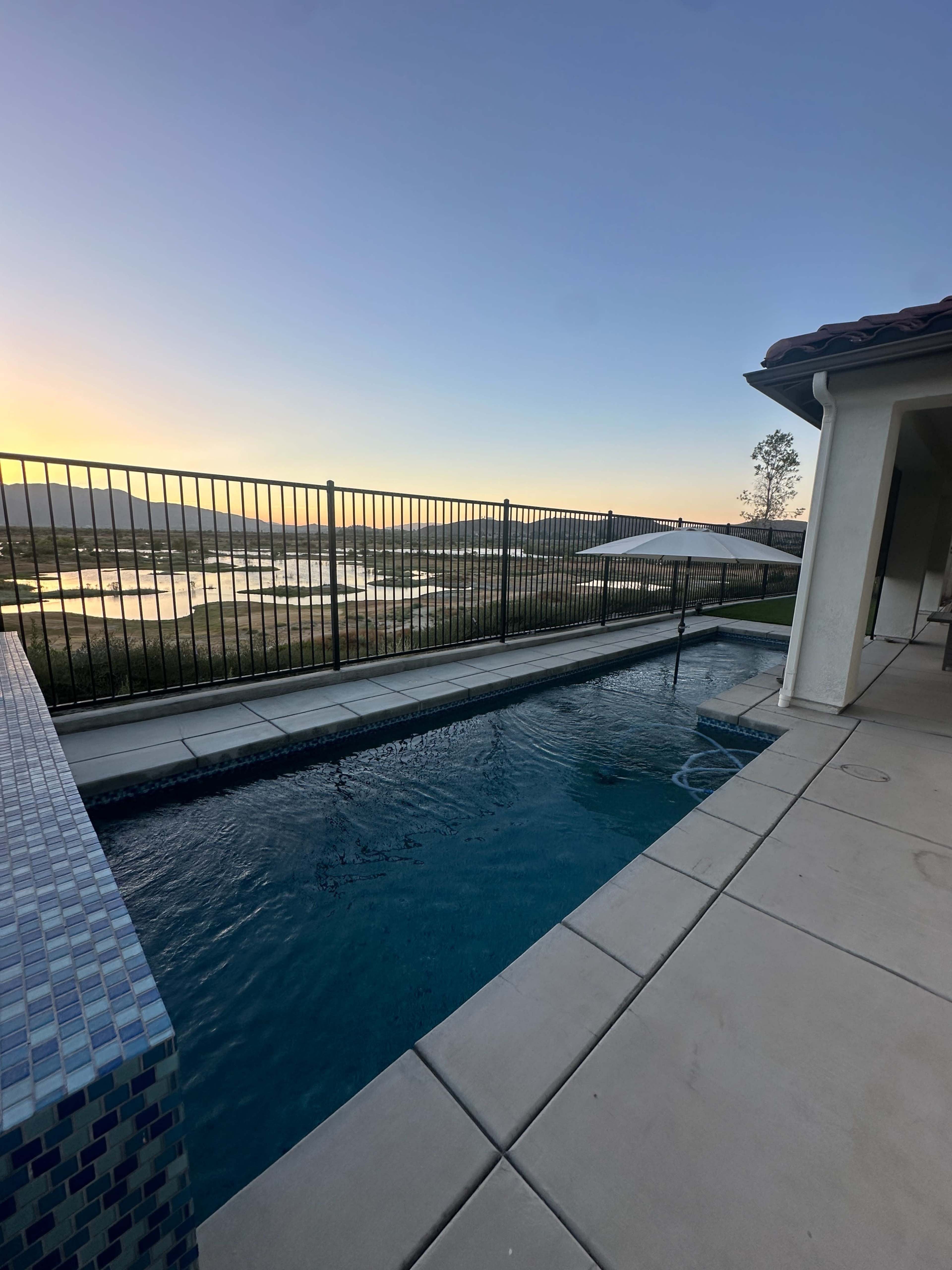 The image shows a swimming pool beside a fenced area overlooking a scenic landscape at sunset.