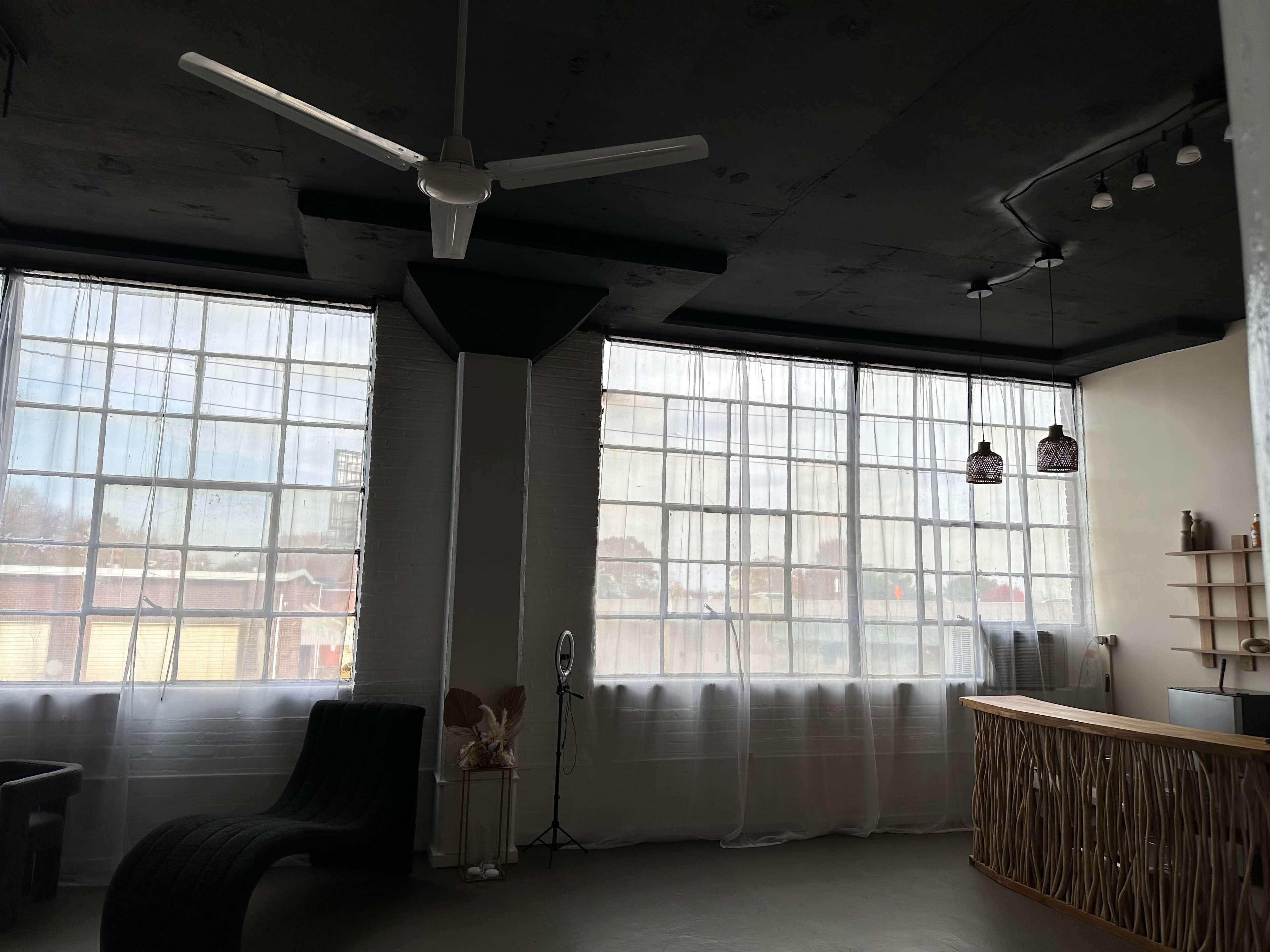 The image shows a spacious room with large windows covered by sheer curtains, a ceiling fan, and a curved seating area, along with a reception desk and decorative elements on the walls.