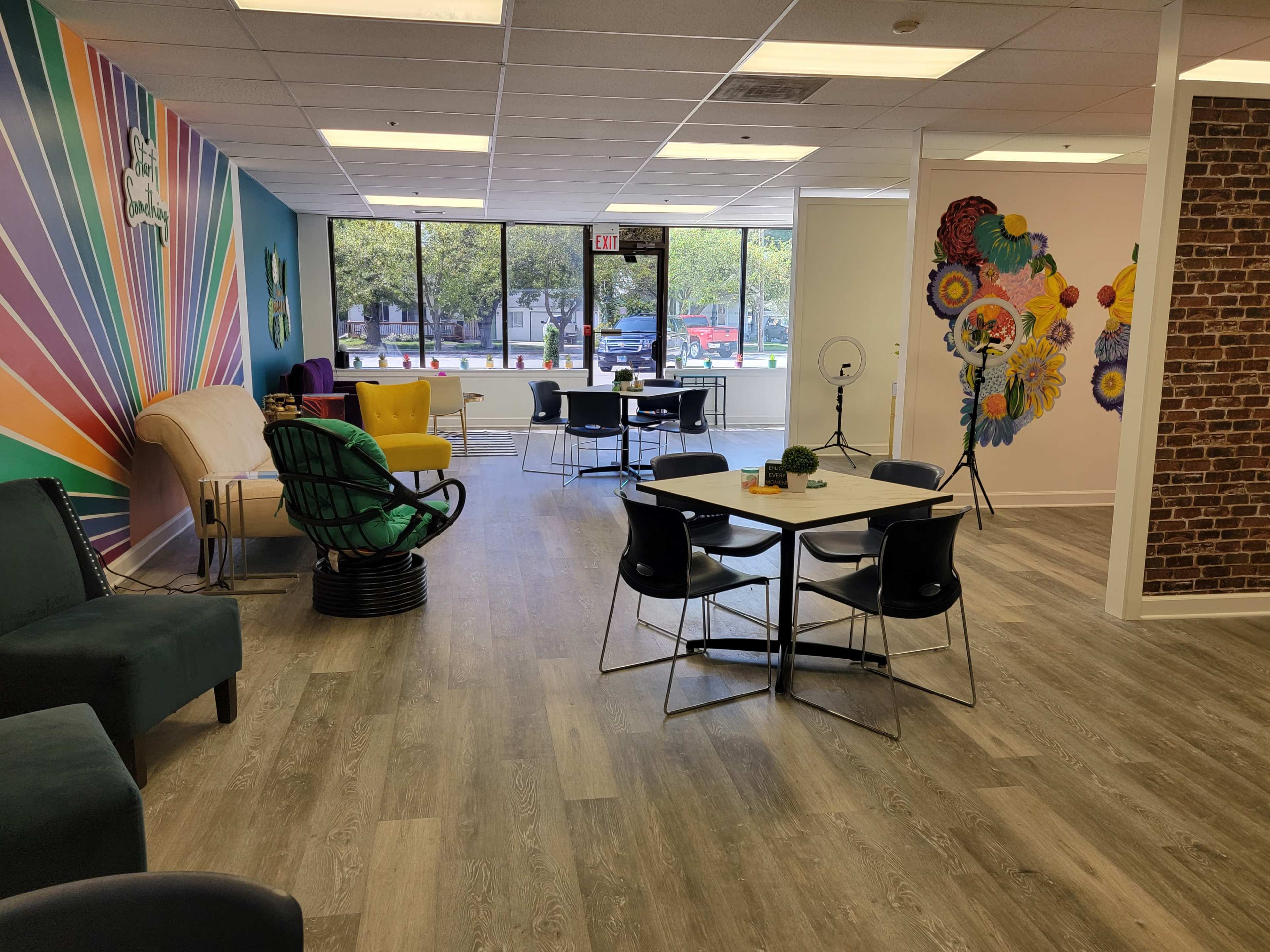 A brightly colored co-working space with modern furniture, including chairs and tables, large windows, and vibrant artwork on the walls.