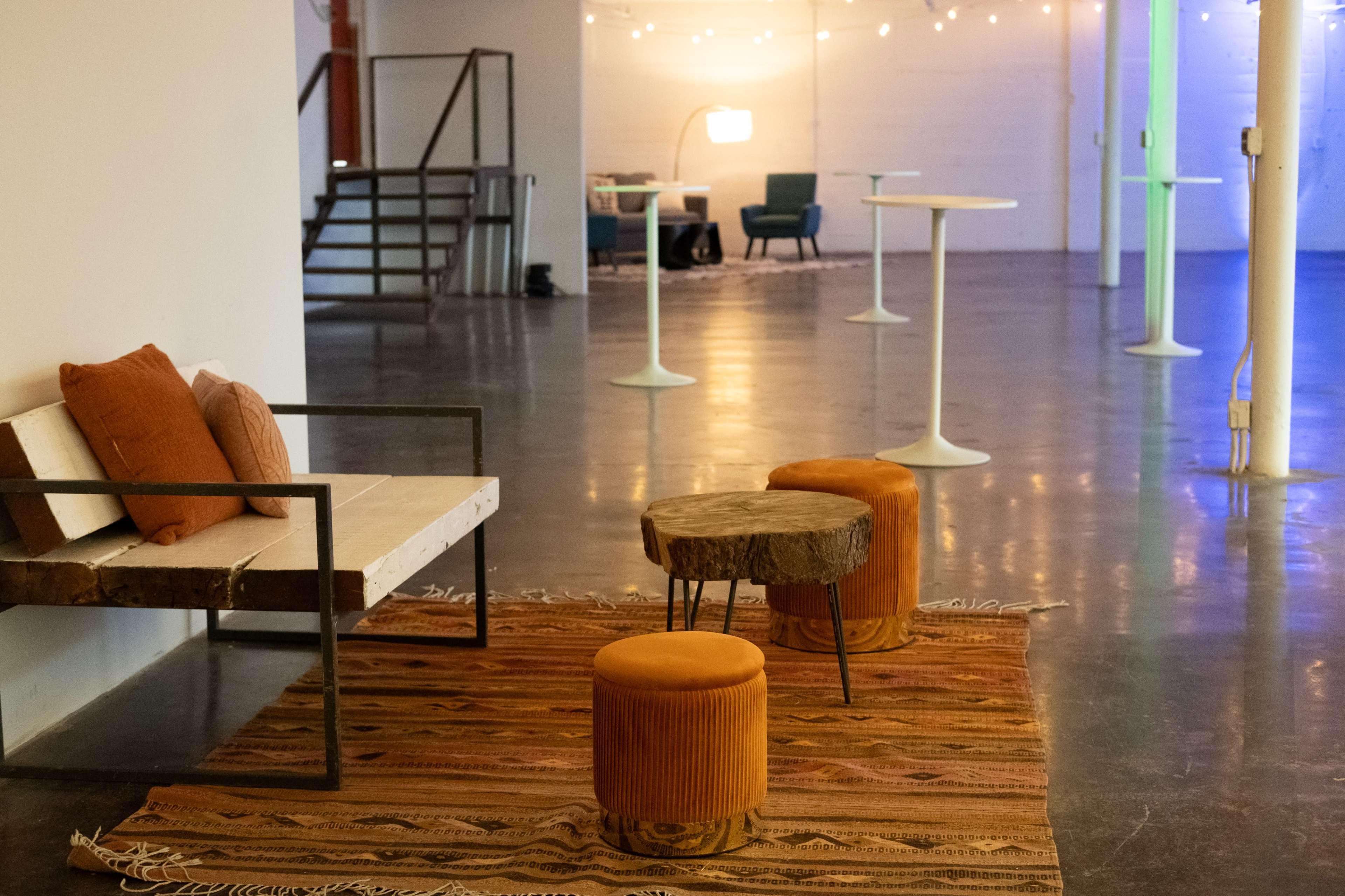 The image shows a modern space with a rustic sofa, two orange stools, and a woven rug, illuminated by ambient lighting.