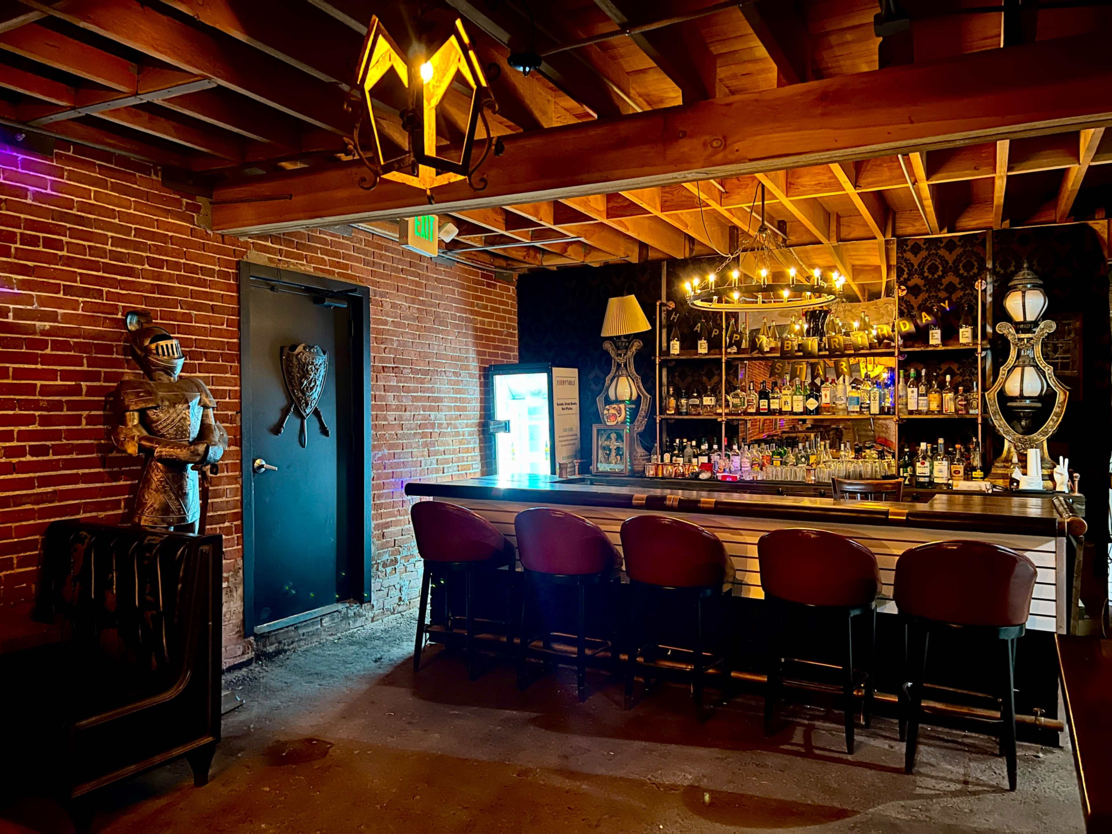 A dimly lit bar area featuring a long counter with several stools, a shelf displaying various bottles behind the bar, and a knight statue by the entrance.
