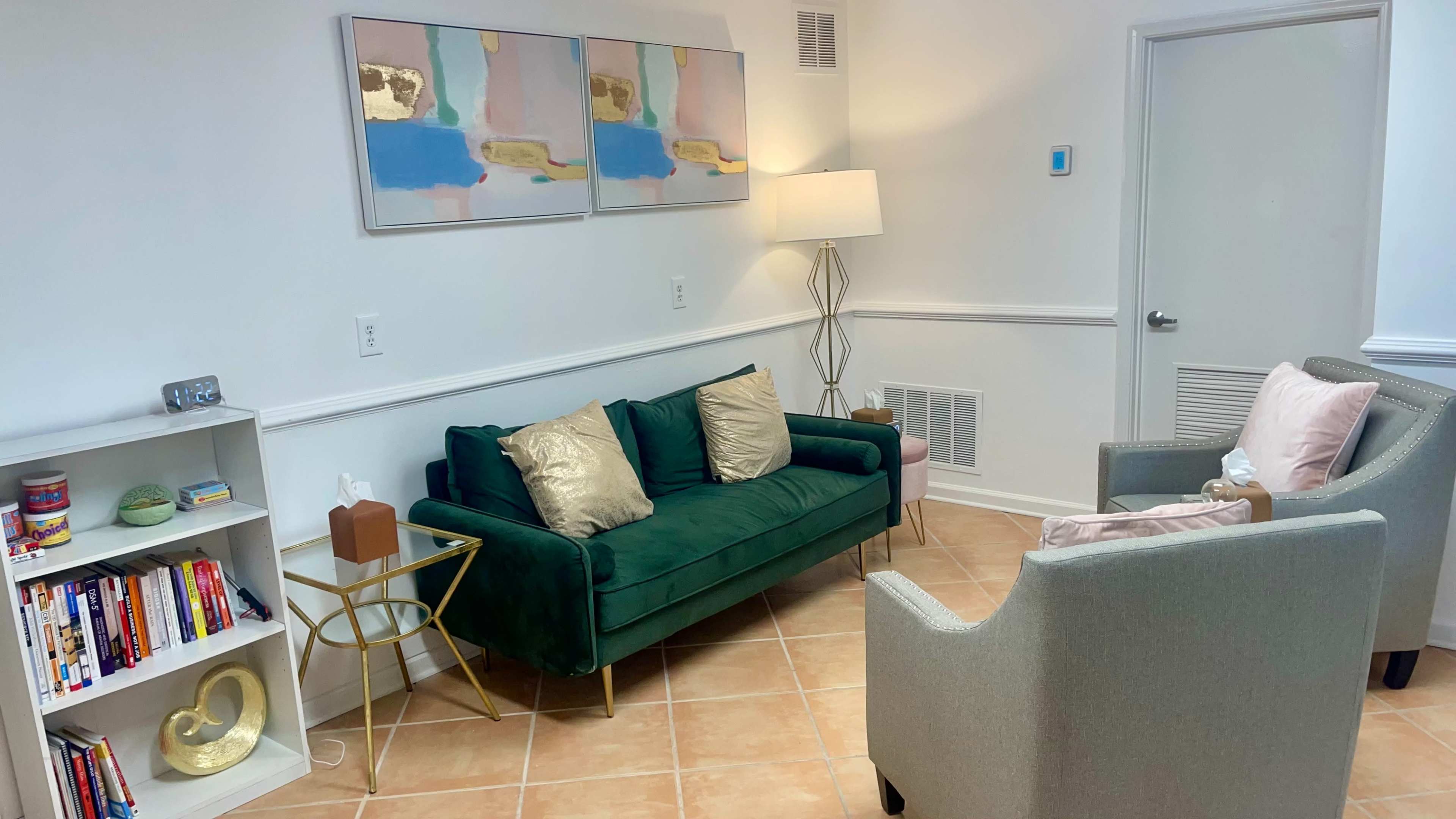 A cozy, well-lit living space featuring a green couch with decorative pillows, two gray armchairs, a gold side table, and a bookshelf filled with books.