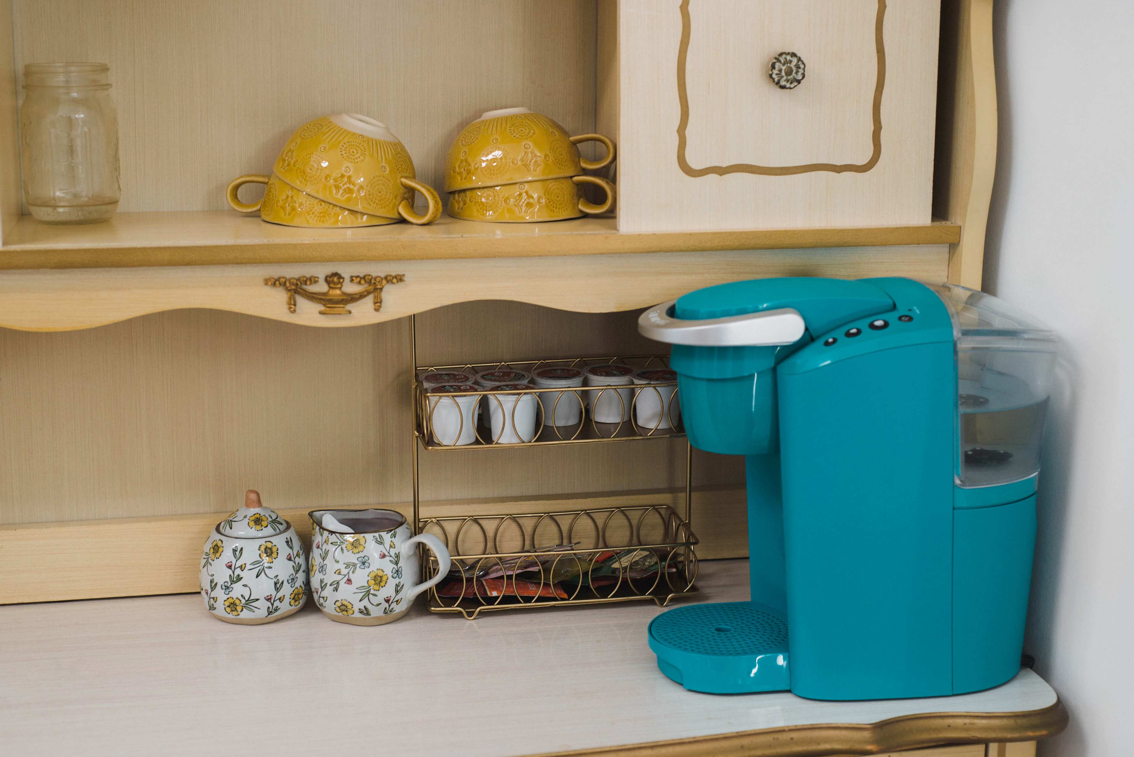 A turquoise coffee maker sits beside a gold and white teacup set on a wooden shelf.
