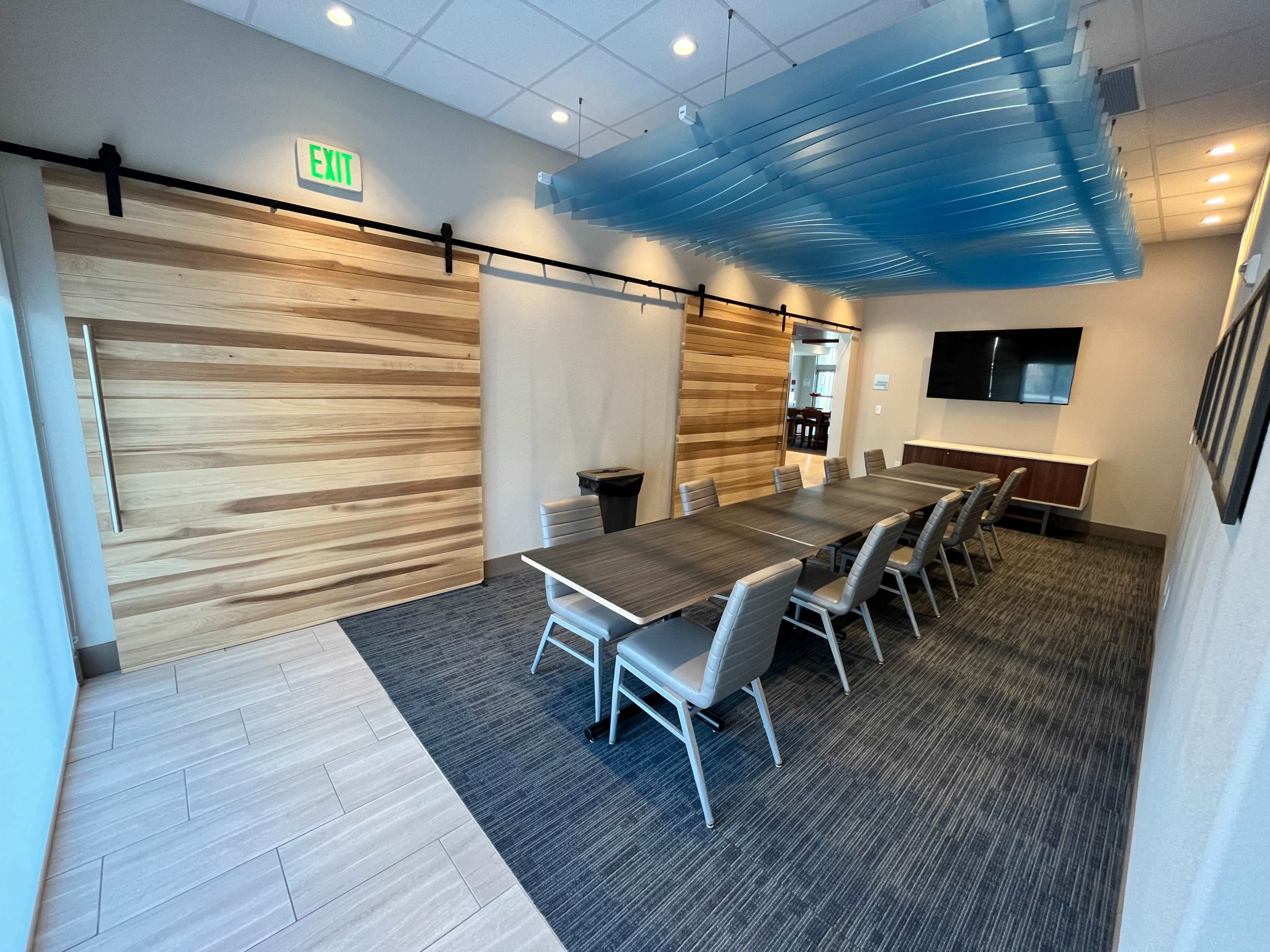 The image depicts a modern conference room featuring a long table with chairs, wooden sliding doors, and a blue ceiling feature.