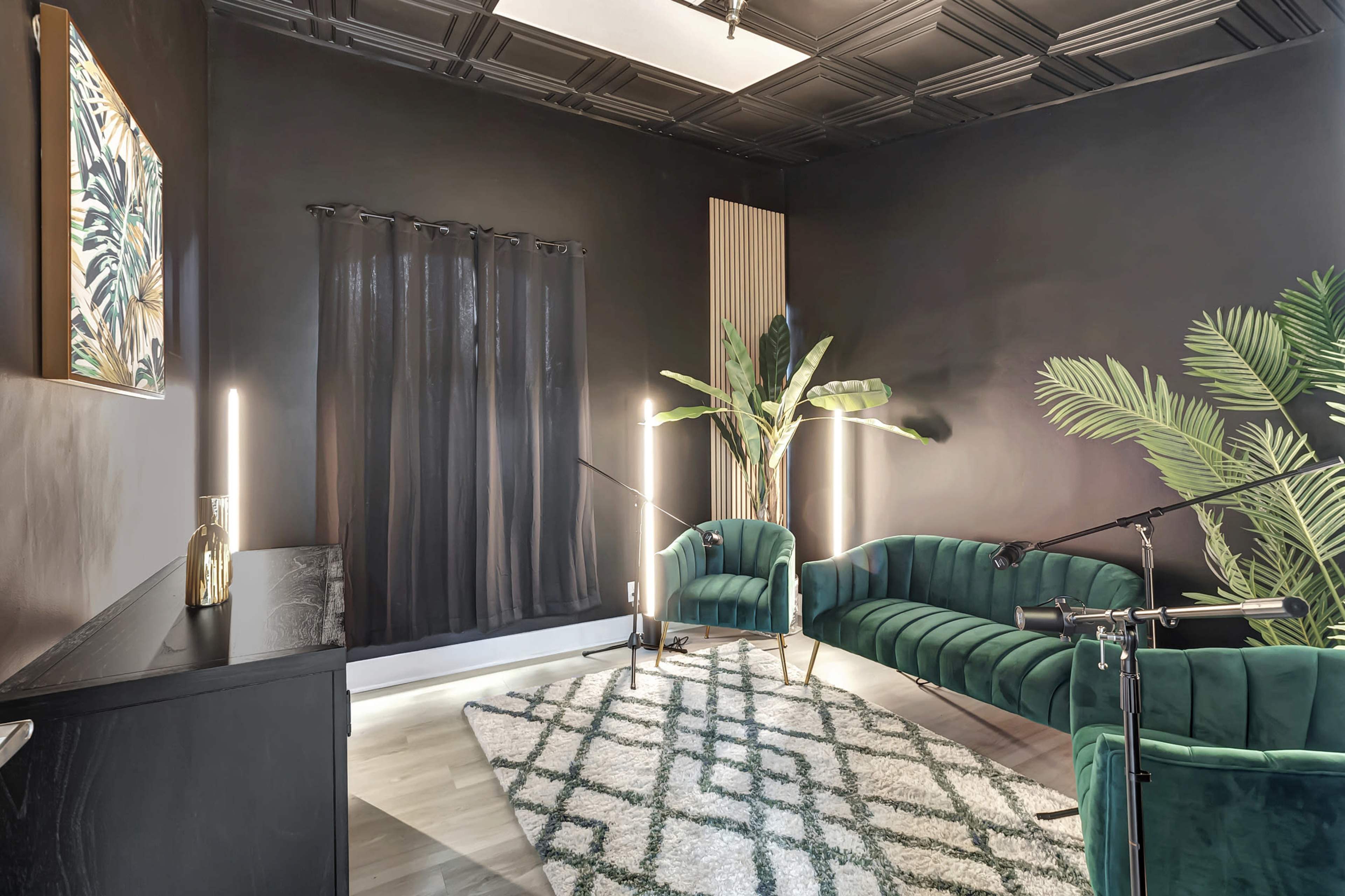 A stylish room featuring two green velvet sofas, a patterned rug, a tall plant, and soft lighting against a dark wall.
