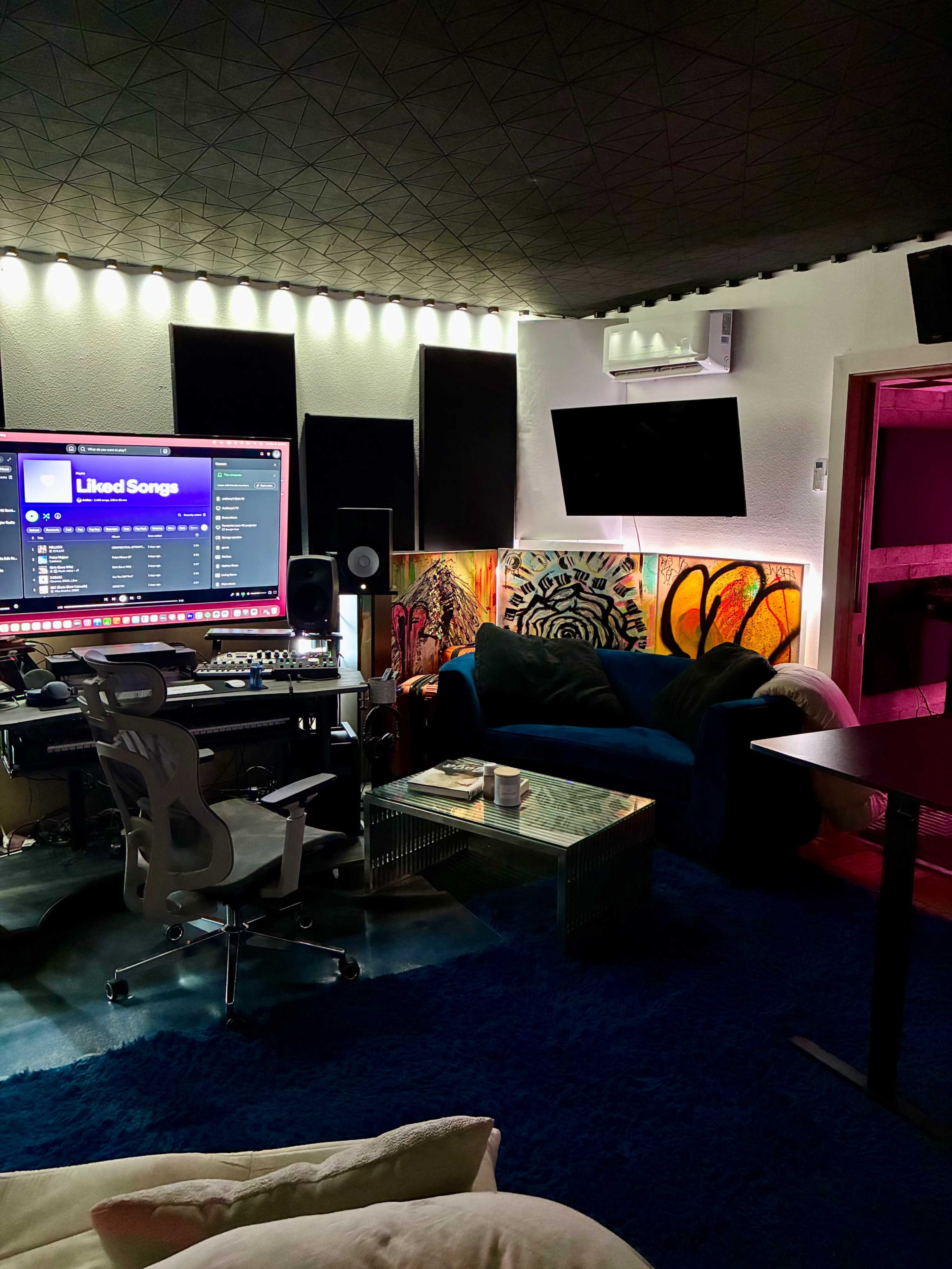 A music studio features a large computer screen displaying a playlist, an office chair, a blue couch, and colorful wall art, with soundproofing panels lining the walls.