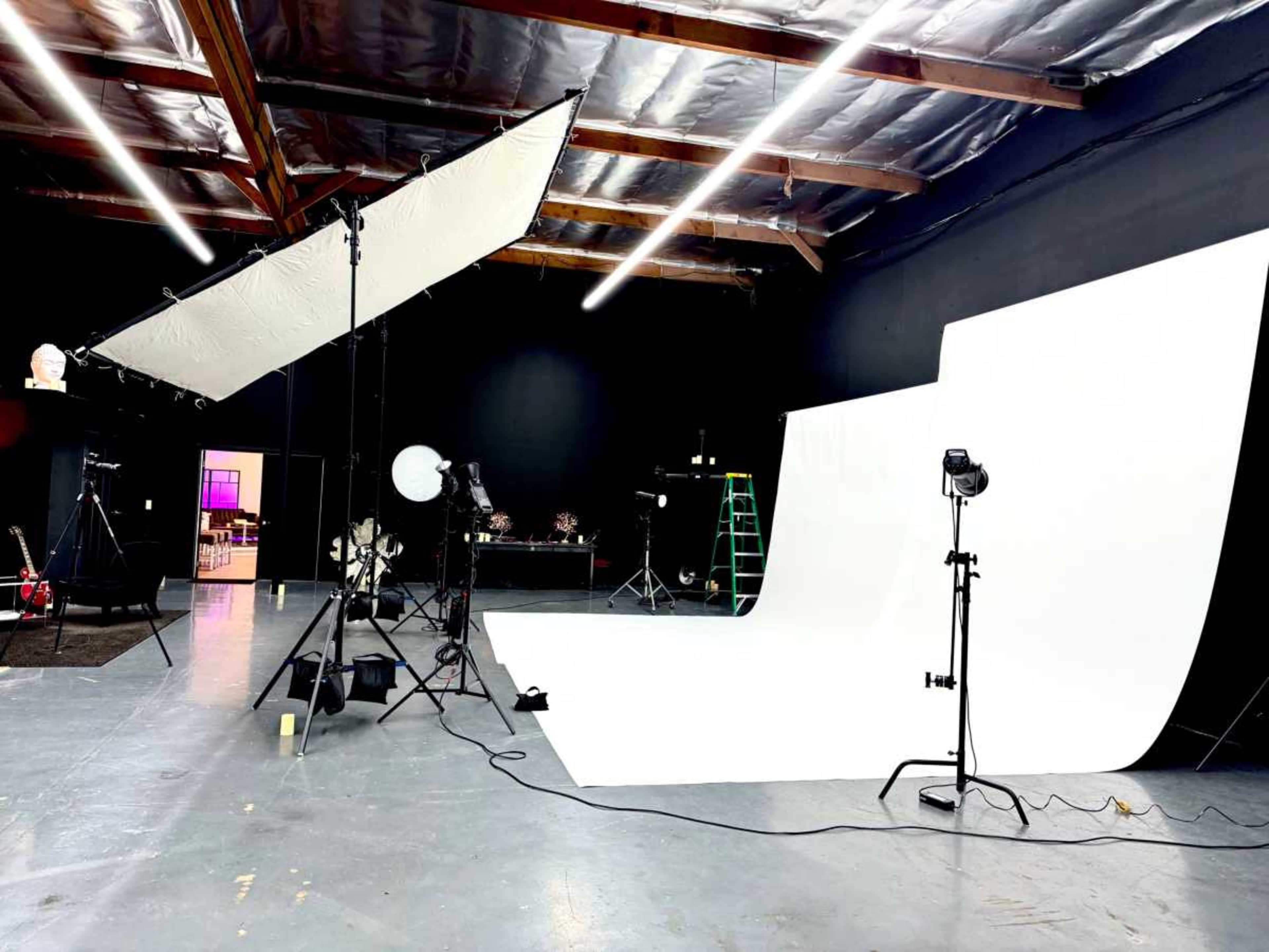 The image shows a photography studio with a large white backdrop, lighting equipment, and various props arranged around the space.