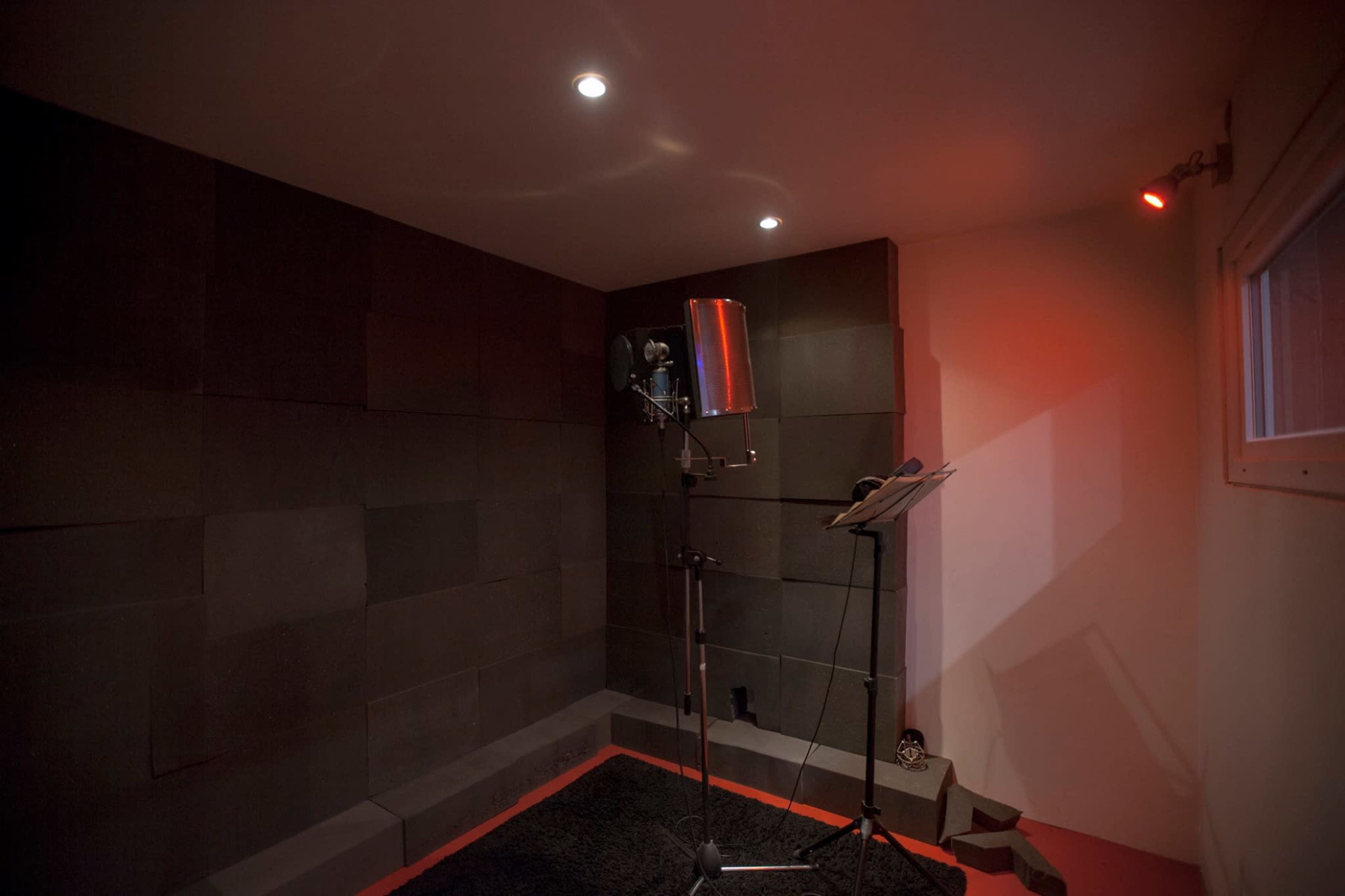 A small soundproof recording studio with dark wall panels, a microphone on a stand, and a music stand in the corner.