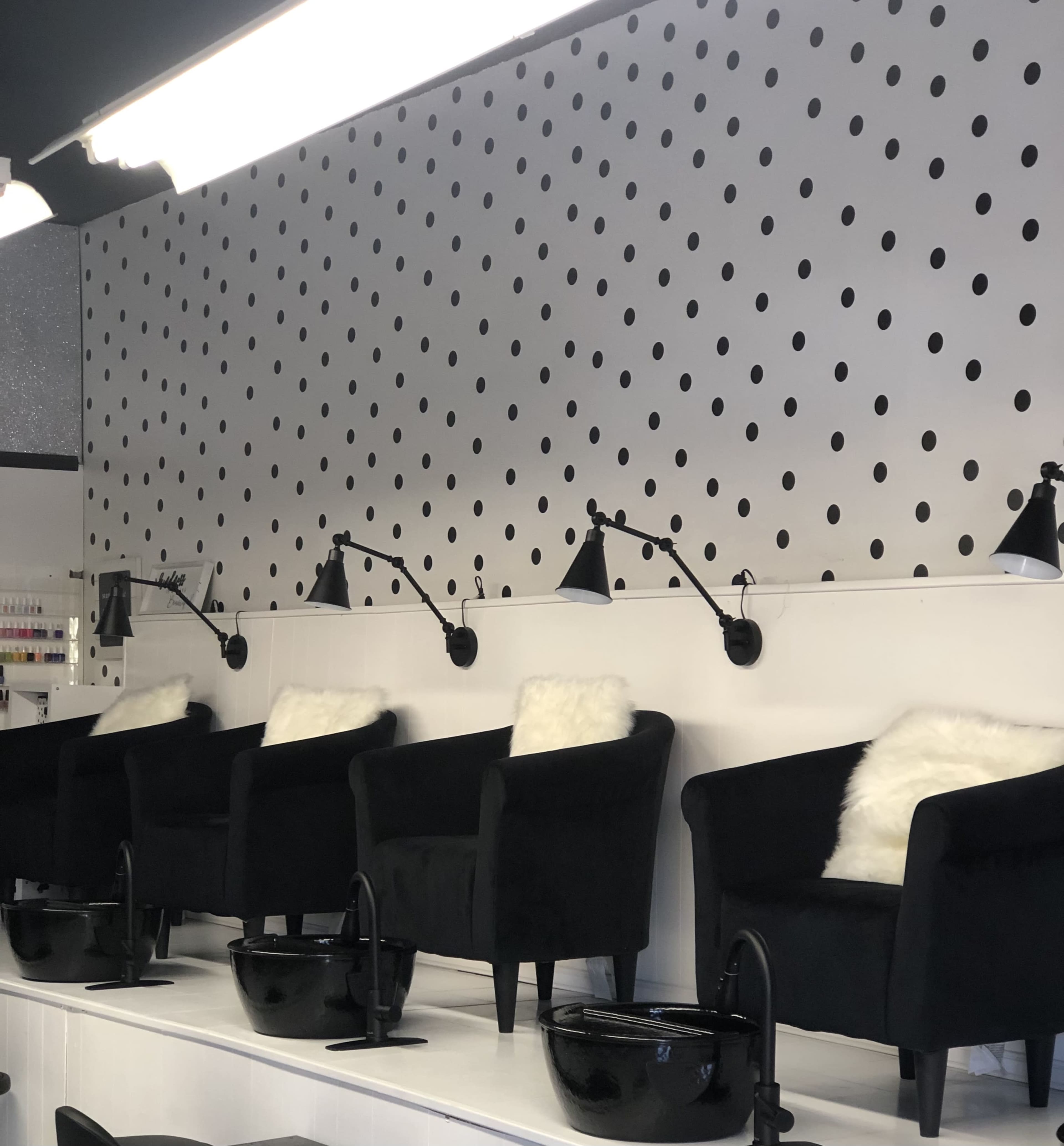 A nail salon interior featuring four black chairs with white fur cushions, positioned in front of black basins against a polka-dotted white wall.