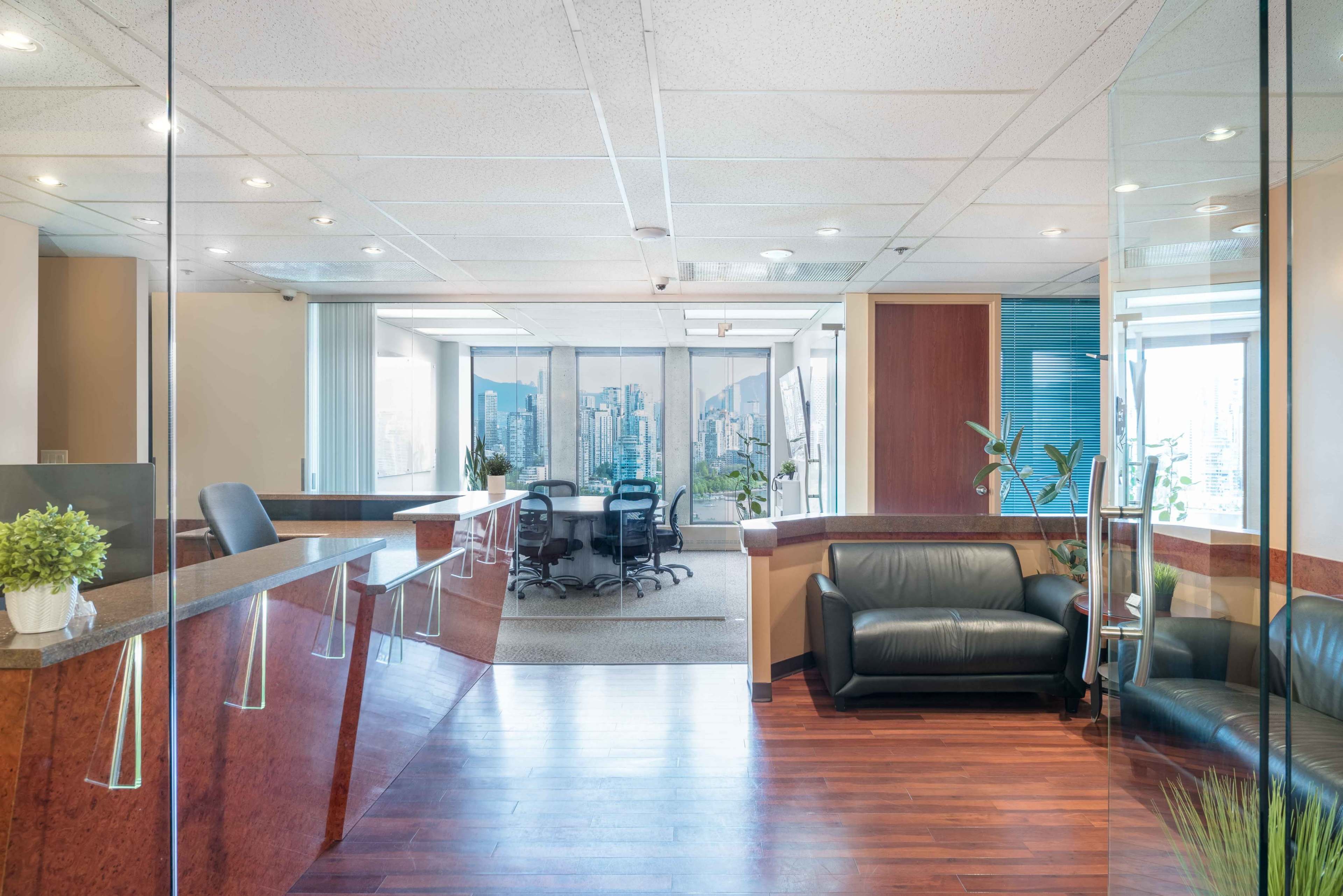 The image shows a modern office reception area with a sleek wooden desk, a leather couch, and a conference room visible through glass walls, overlooking a city skyline.