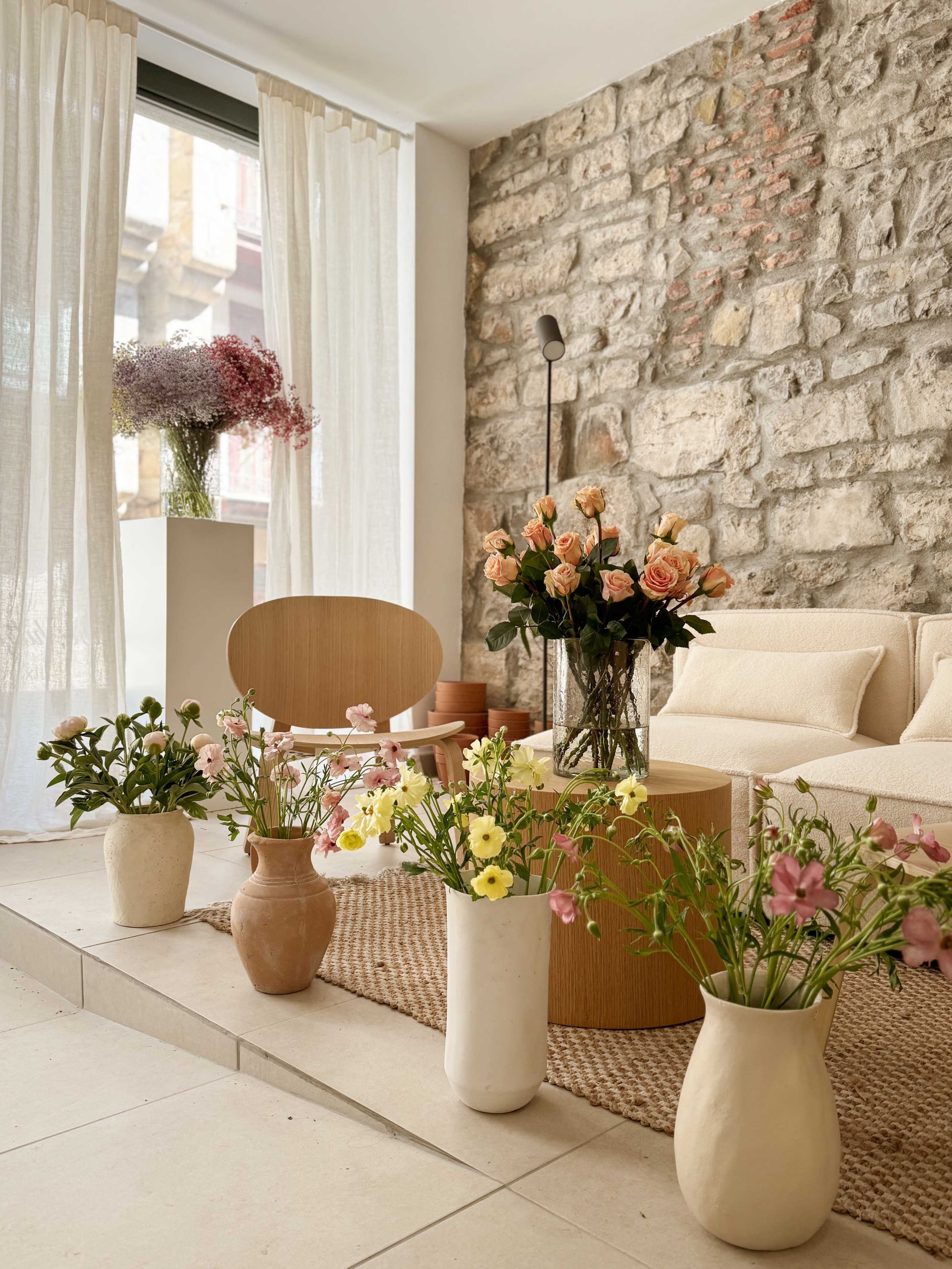 The scene features a modern living room with a stone wall, a beige sofa, a wooden chair, and various flower arrangements in vases on the floor.