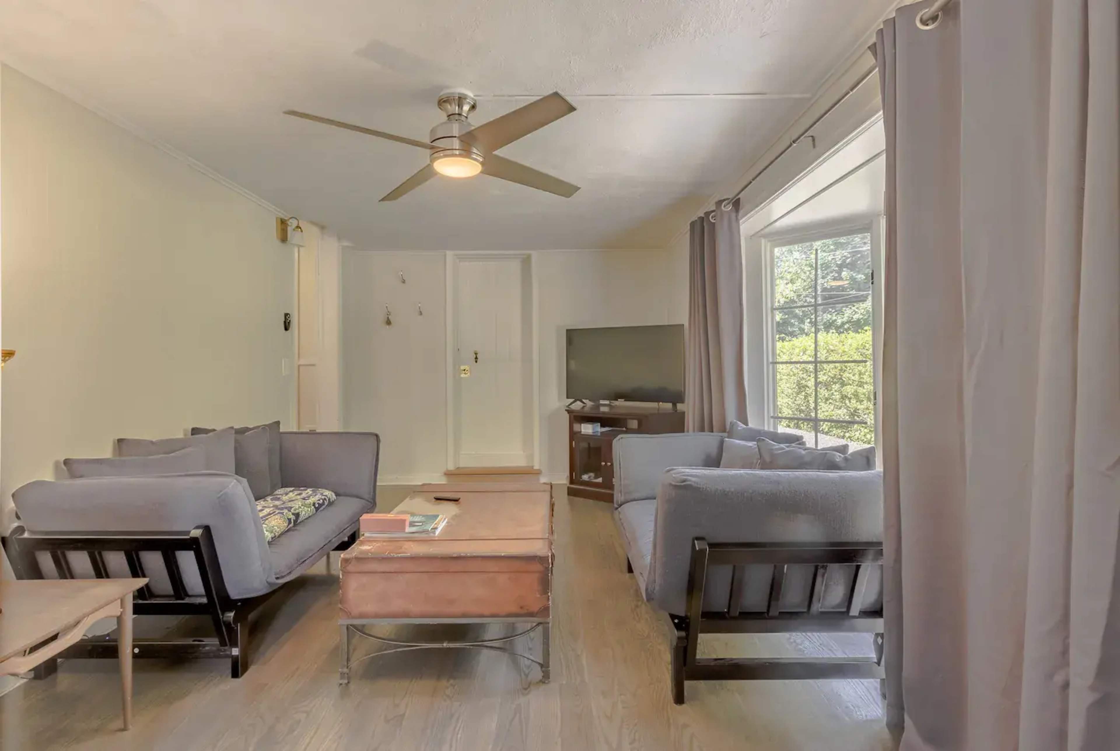 A living room features two gray couches, a wooden coffee table, a television, and large windows with natural light streaming in.