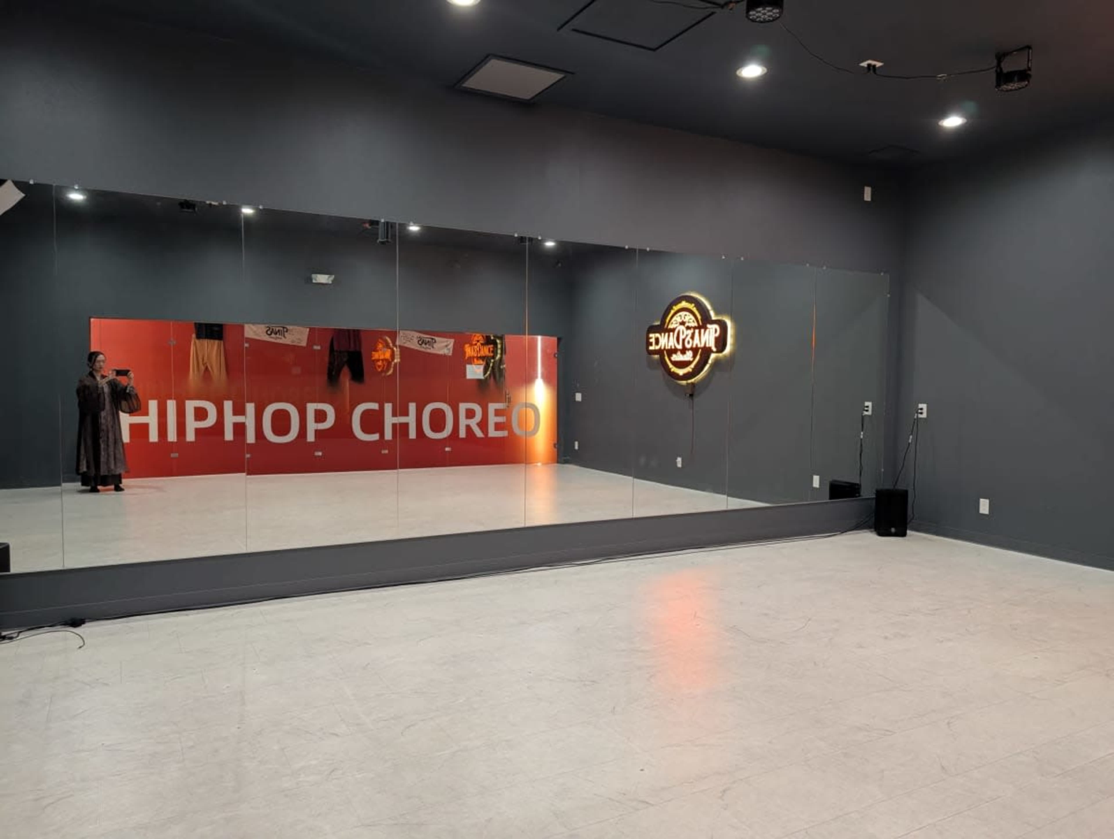 The image shows a dance studio with mirrors on one wall and a neon sign that reads "HIPHOP CHOREO."