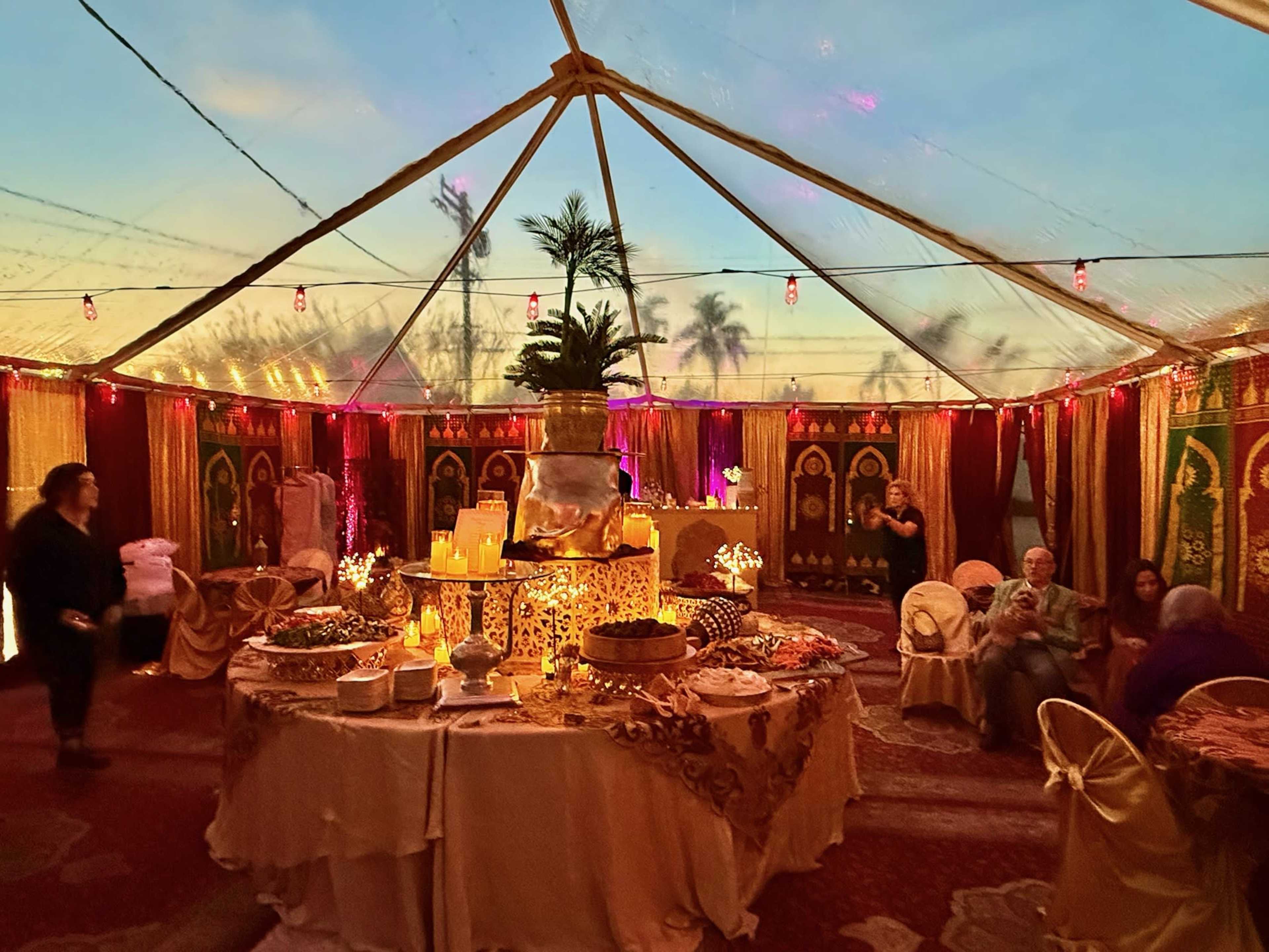 A spacious tent is decorated with colorful fabrics and lights, featuring a table laden with various dishes and surrounded by guests in a festive setting.