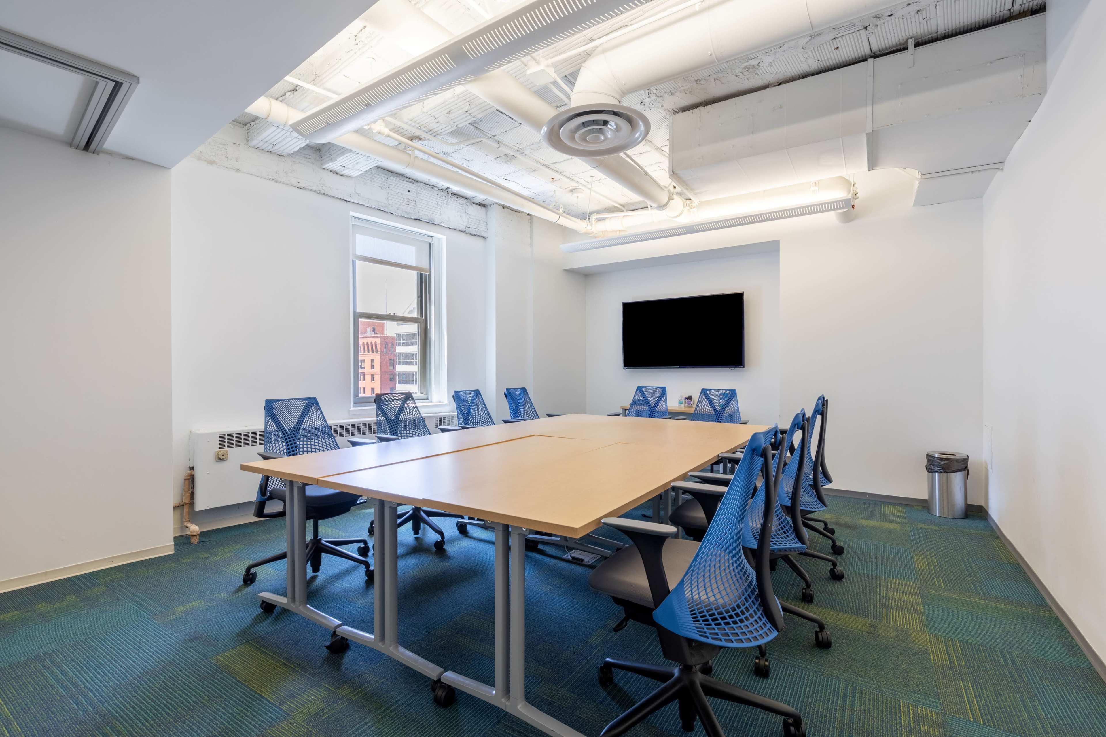 A modern conference room features a long wooden table surrounded by blue office chairs, with a wall-mounted screen and large windows allowing natural light.